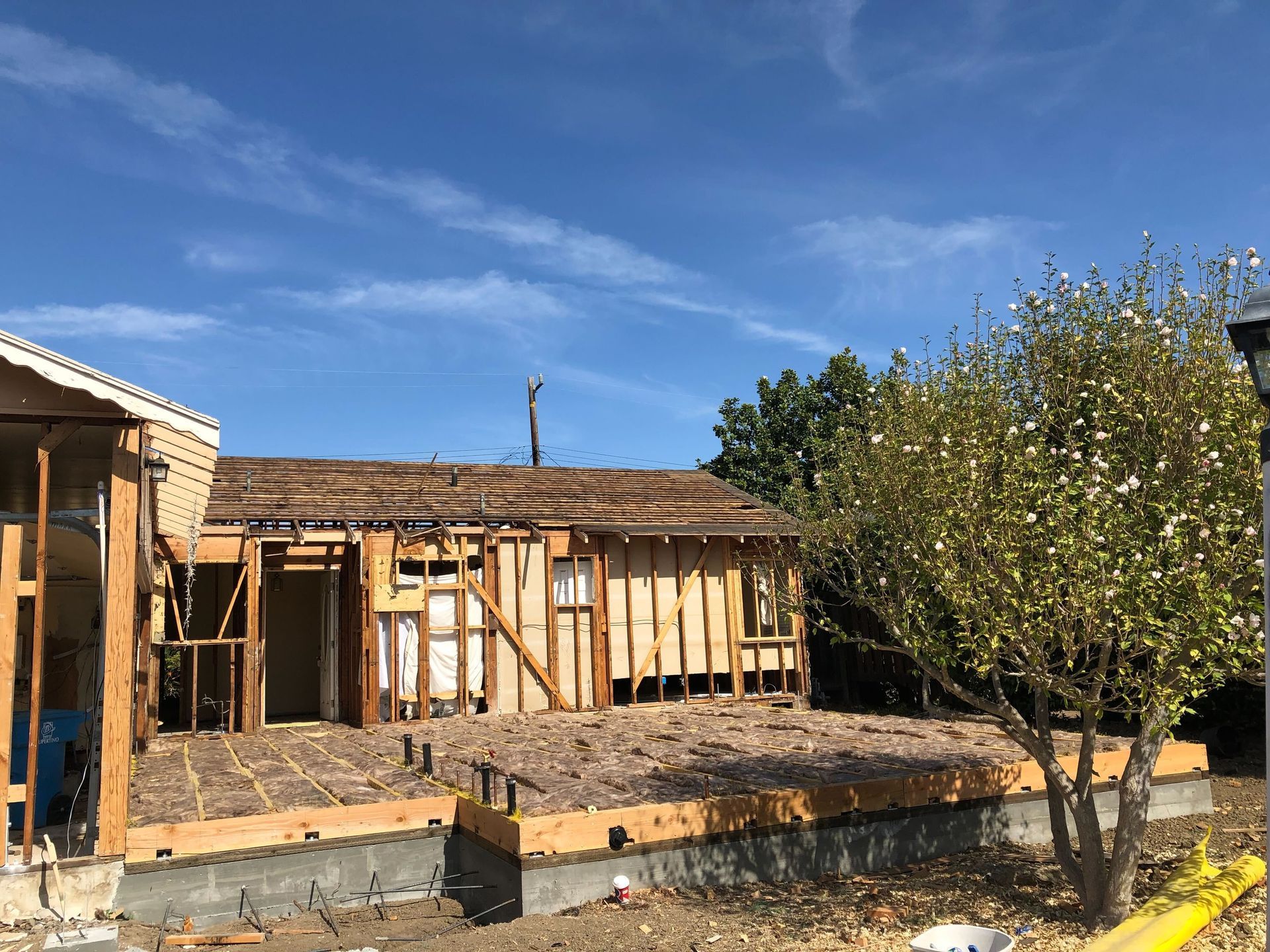 Partially built wooden house under construction beside a dirt yard and tree under a blue sky