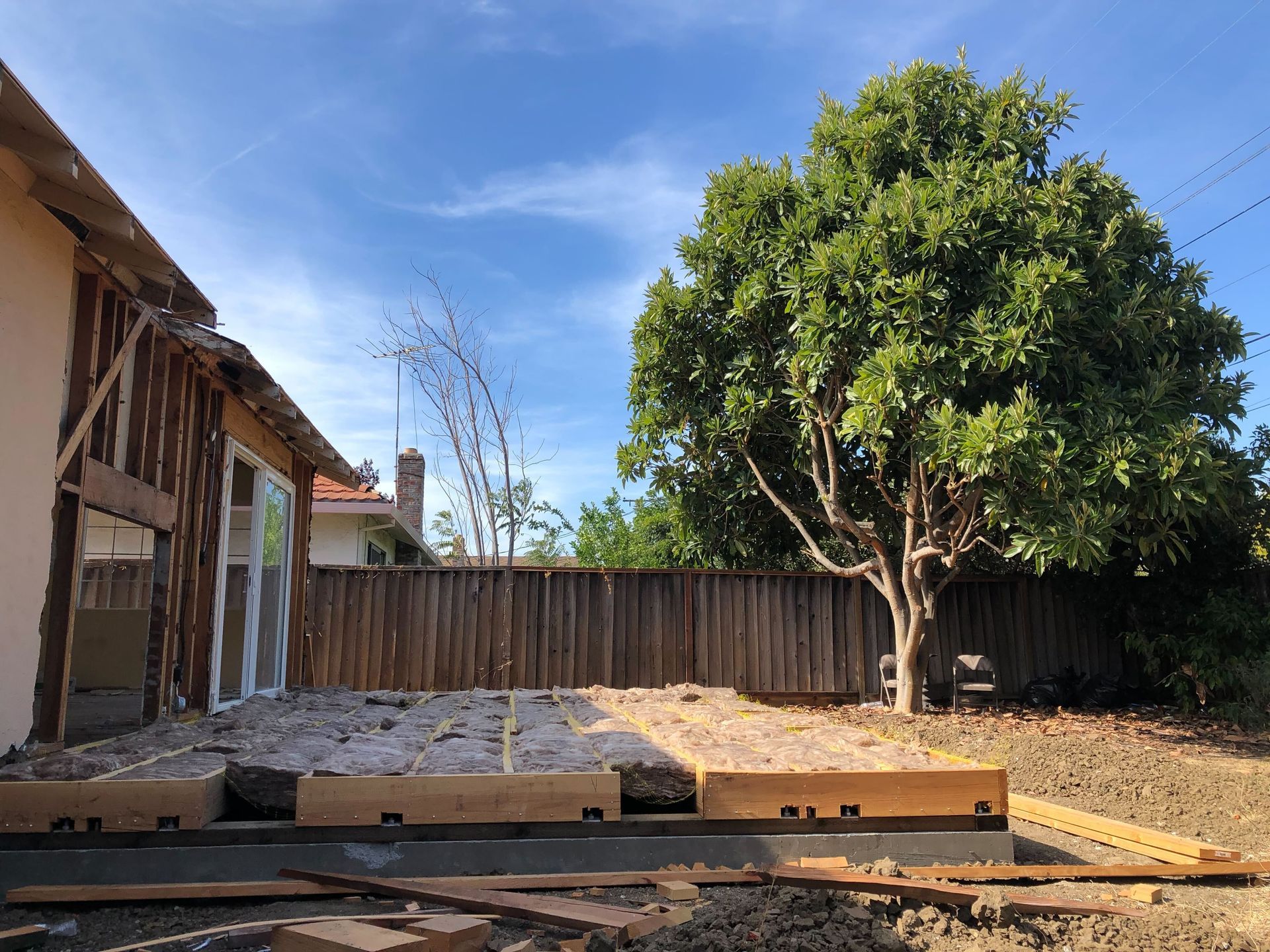 Backyard construction site with wooden framing, concrete slab, and a large tree under a blue sky