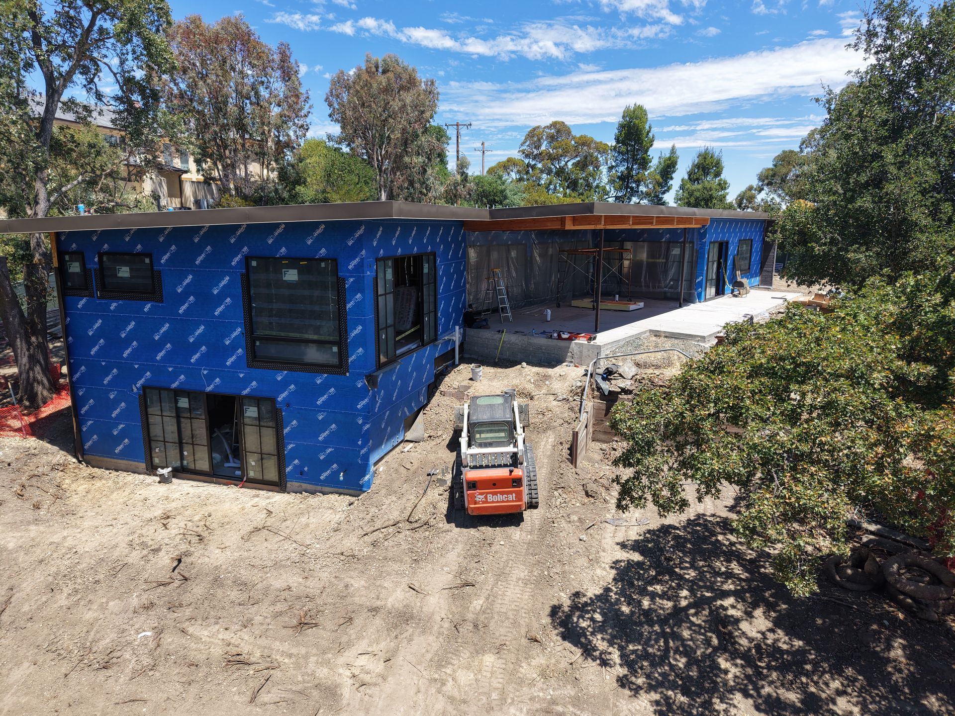 Blue house under construction with a red skid steer in front and trees around it