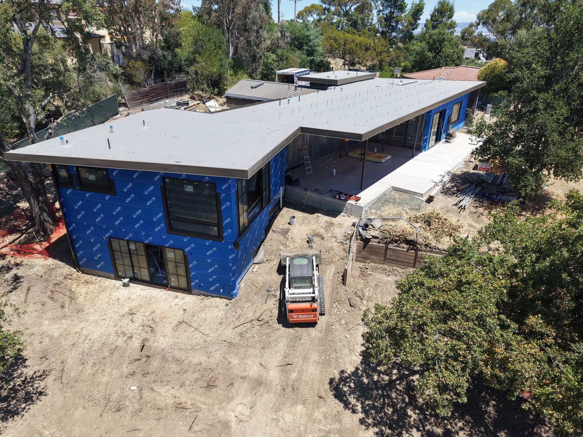 Aerial view of a blue building with a gray roof, surrounded by trees and dirt, with a small vehicle nearby