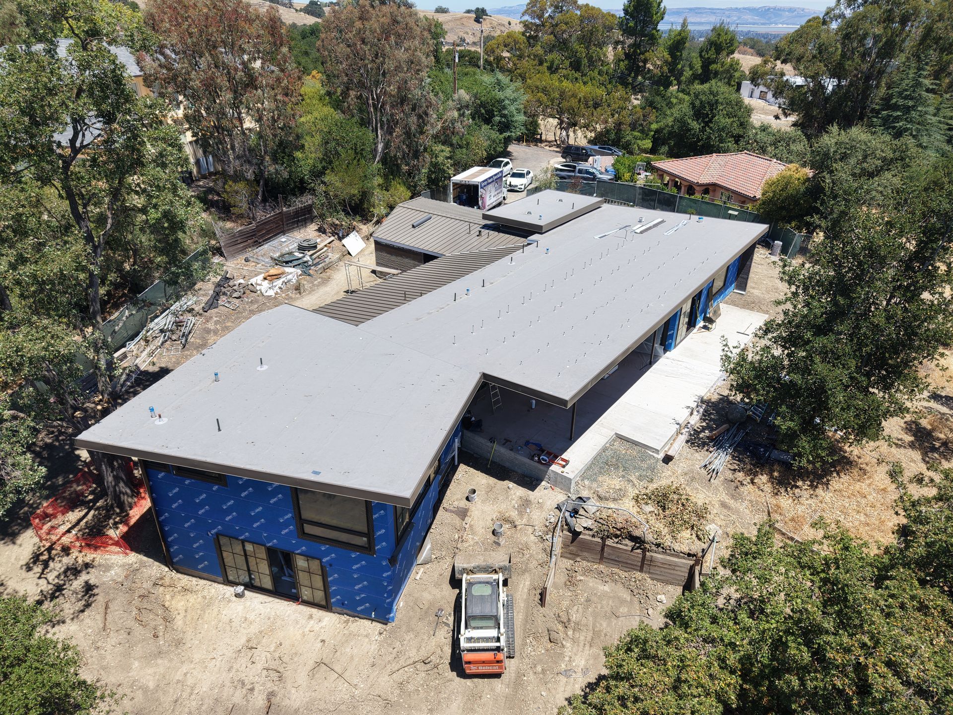 Aerial view of a blue building with a gray roof under construction, surrounded by trees and dirt.