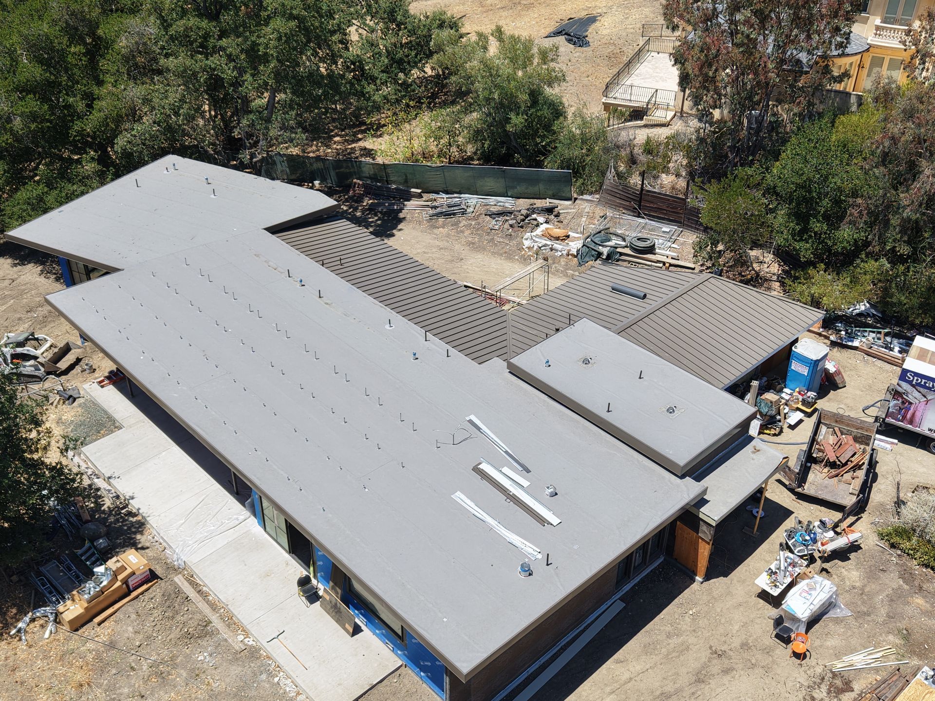 Aerial view of a house with a gray roof and surrounding construction materials in a wooded area