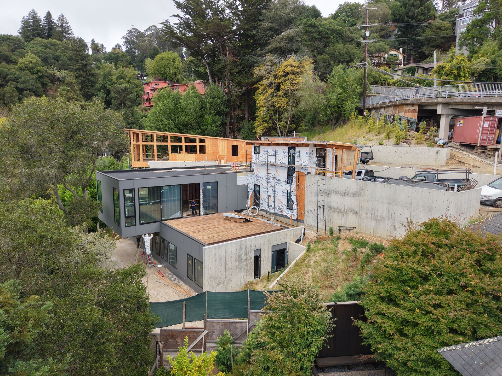 Modern house under construction on a wooded hillside, with exposed concrete, glass, and orange framing.