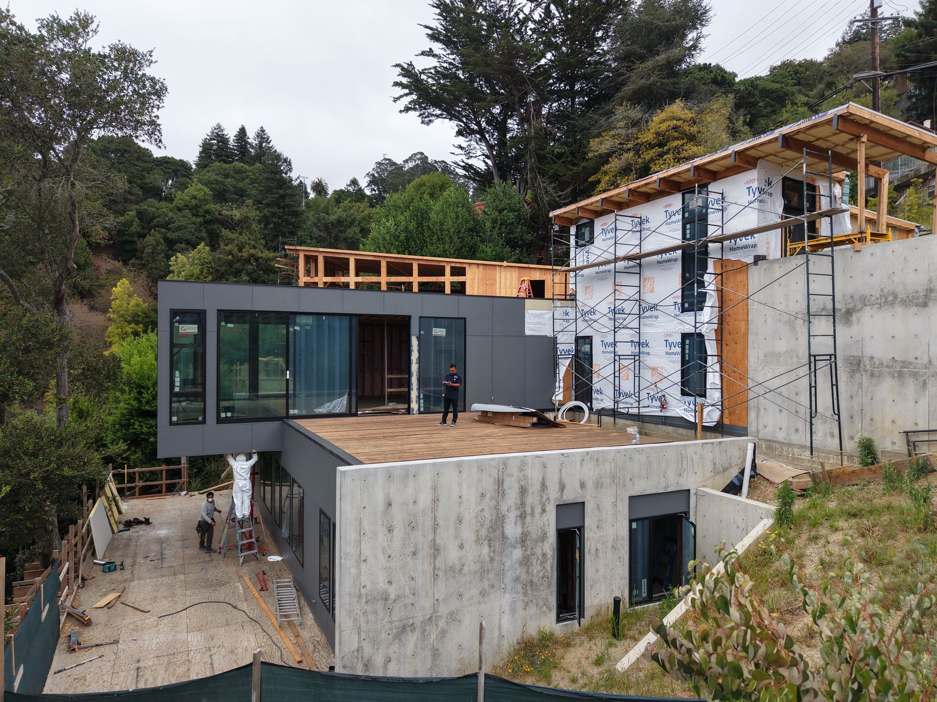 Modern house under construction on a hillside, with concrete walls, dark windows, and surrounding trees.
