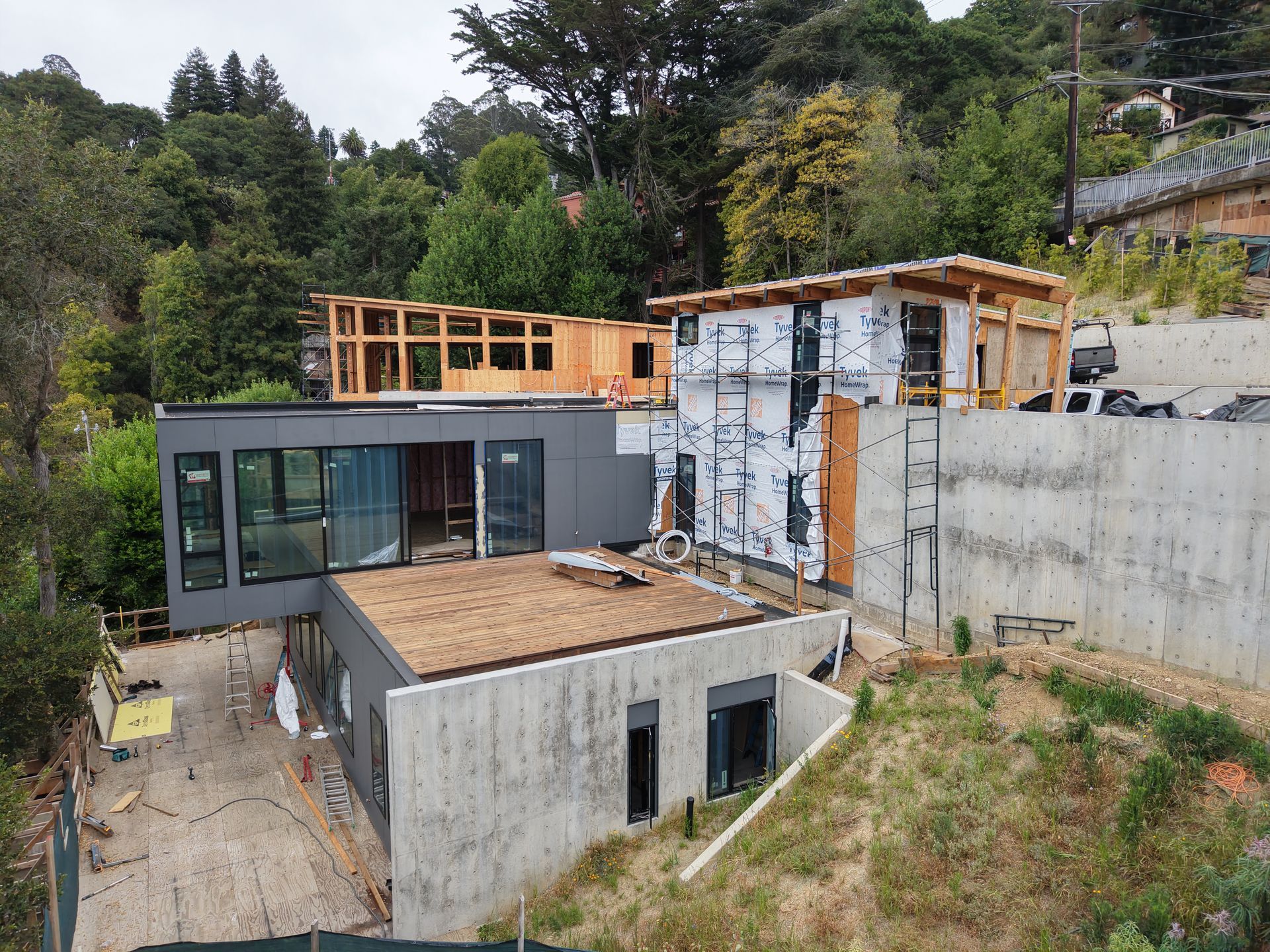 Modern house under construction on a hillside, with concrete walls, unfinished roofs, and surrounding trees
