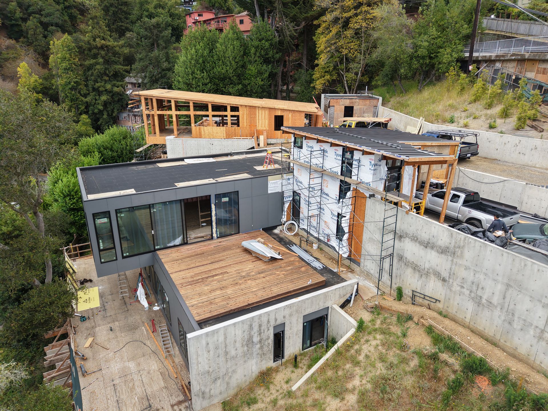 Modern house under construction on a hillside, with concrete walls, wooden framing, and surrounding trees.