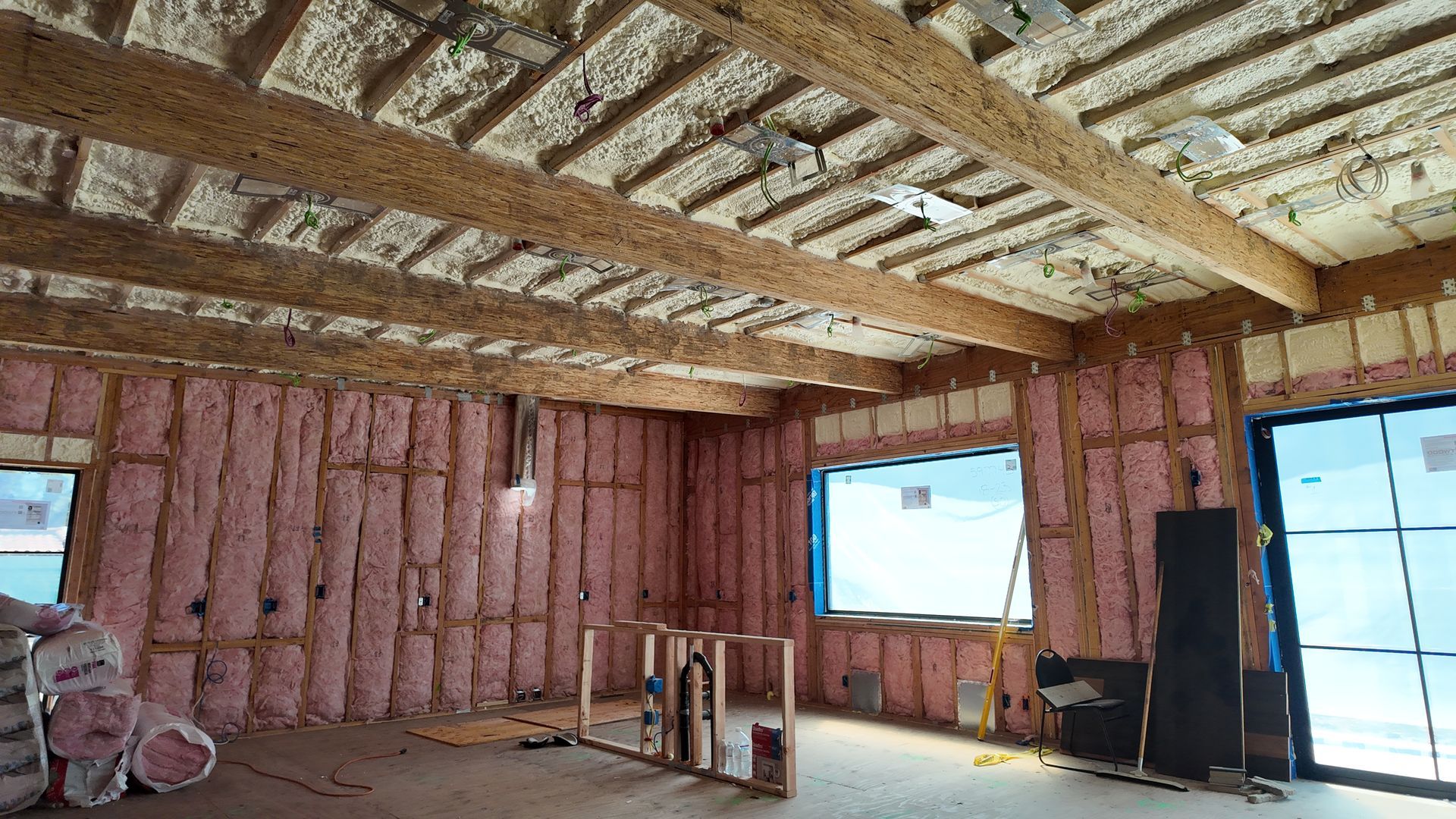 Unfinished room with exposed insulation, wooden ceiling beams, windows, and construction materials inside.