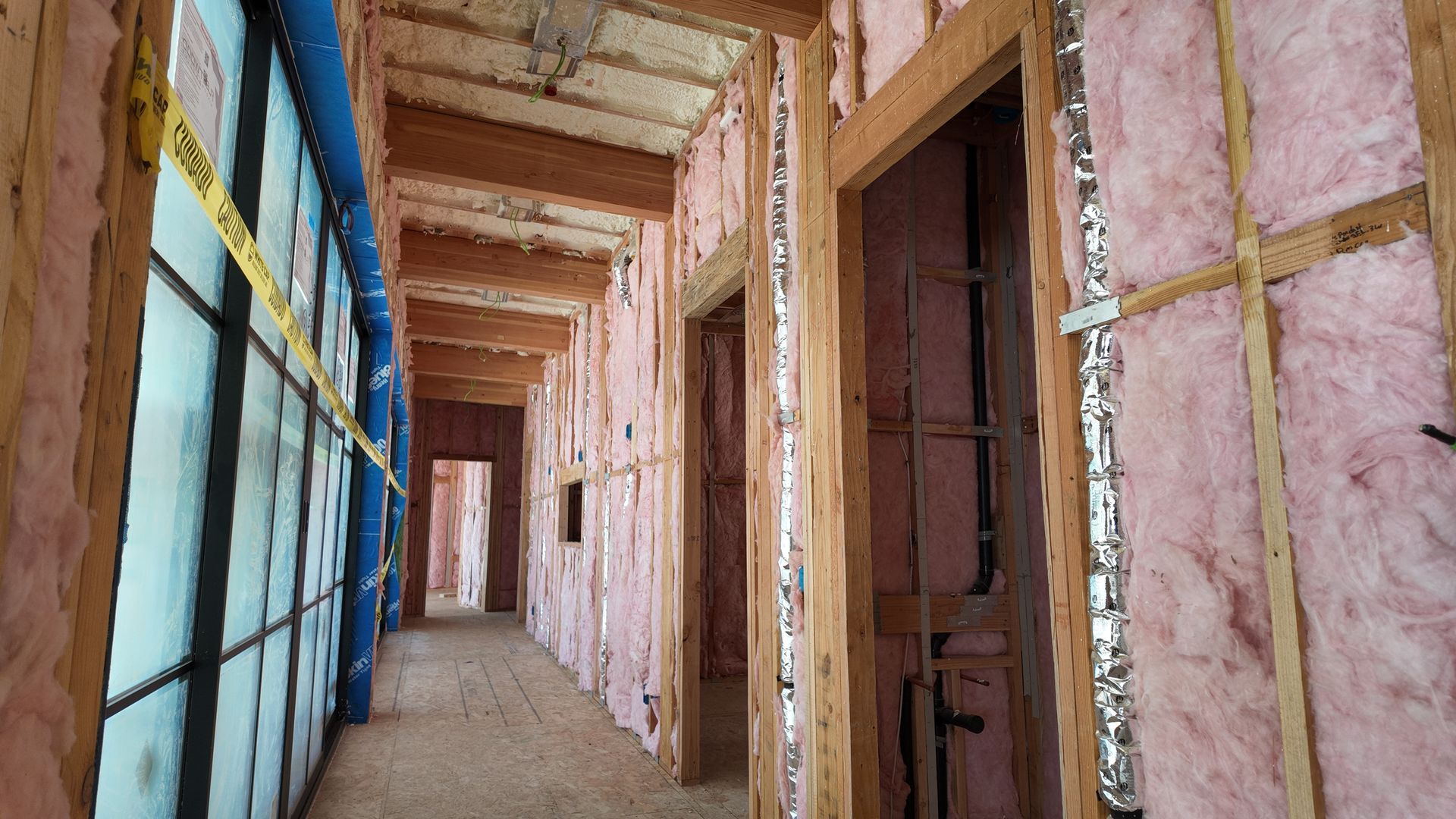 Long corridor under construction with exposed pink insulation, wooden framing, and blue plastic sheeting