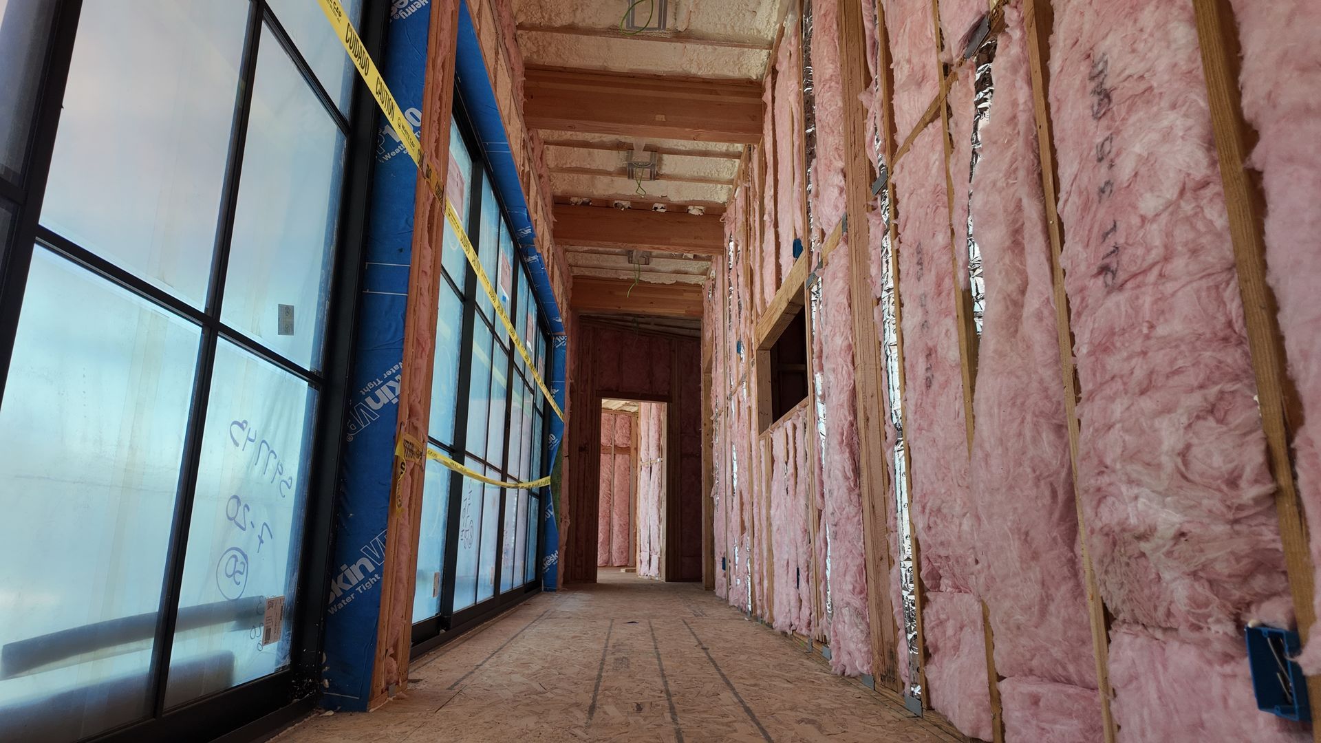 Narrow unfinished hallway with pink insulation walls and large blue-framed windows on the left