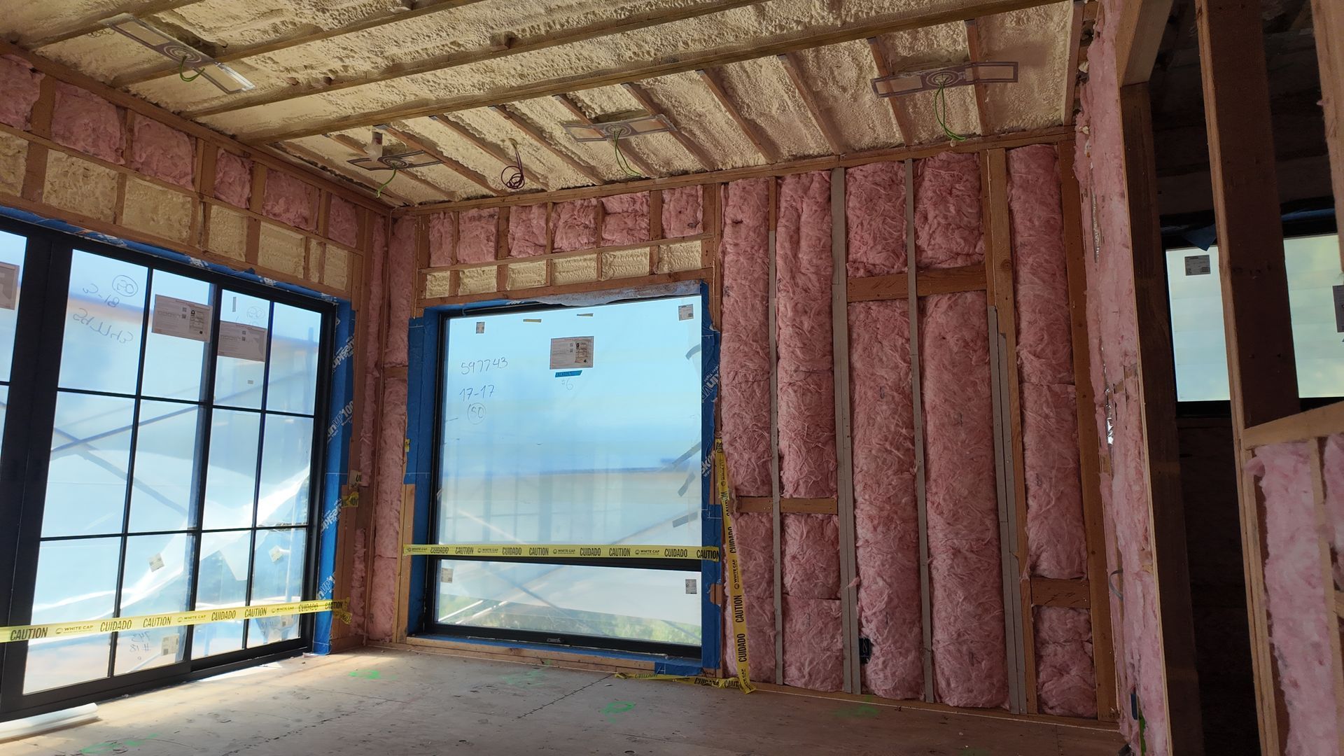 Interior of a house under construction with pink insulation, exposed framing, and large windows.