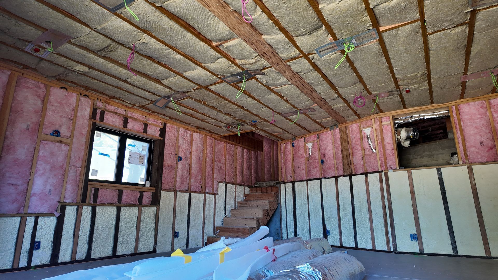 Partially renovated room with exposed insulation, window, stairs, and an open wall cavity