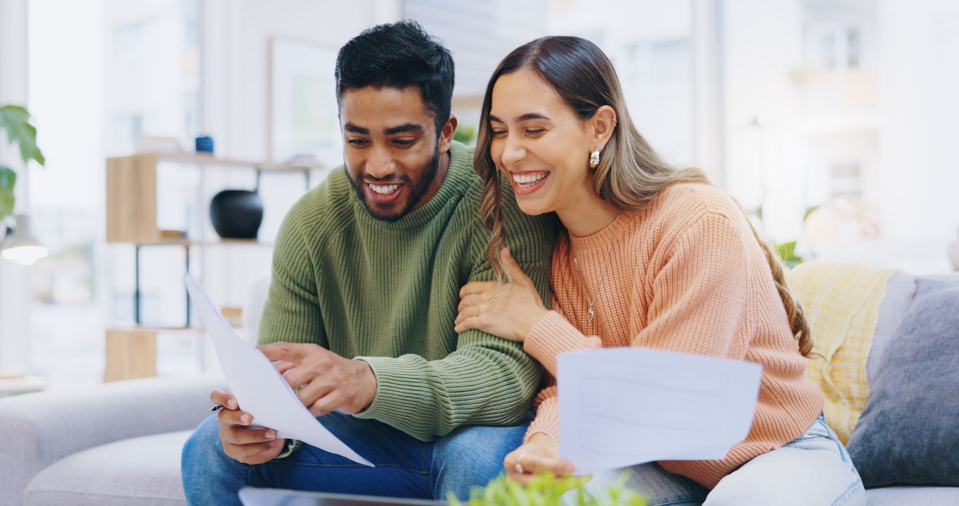 Two people smiling on a couch, reading papers together in a bright living room.