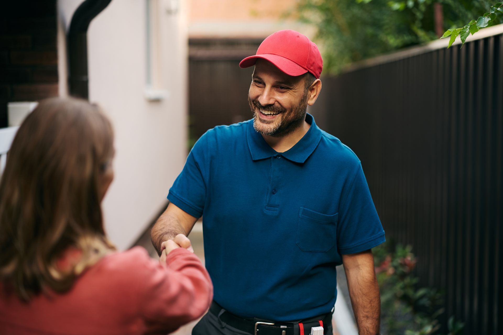Smiling man in red cap shakes hands with a person outdoors by a fence.