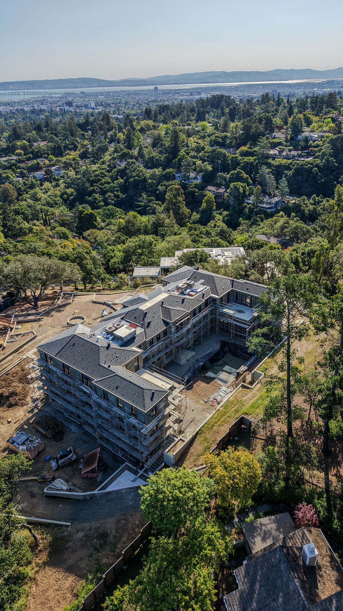 Aerial view of a large building under construction surrounded by dense green trees and a distant cityscape.