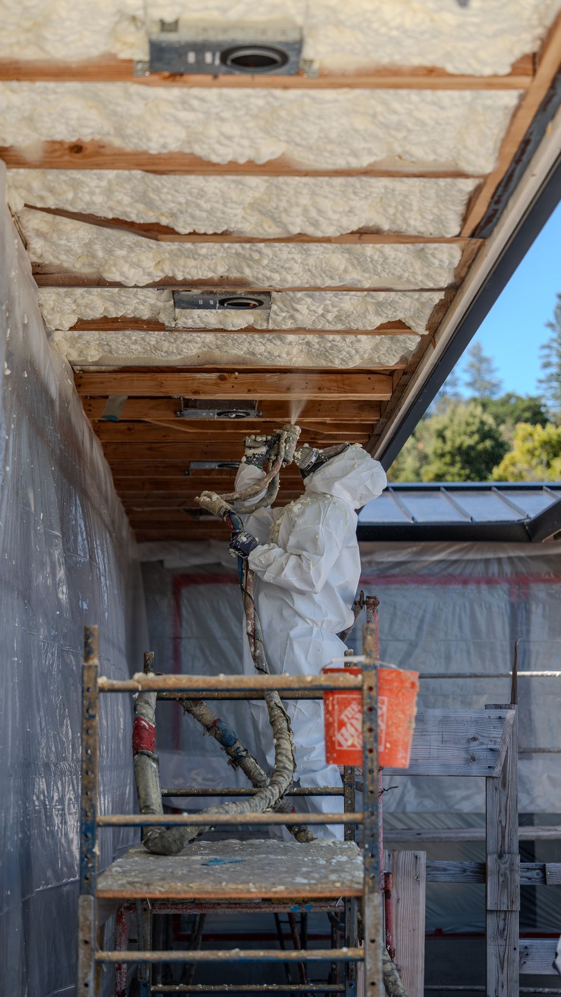 Worker in white coveralls on scaffolding, removing insulation from a ceiling under construction.