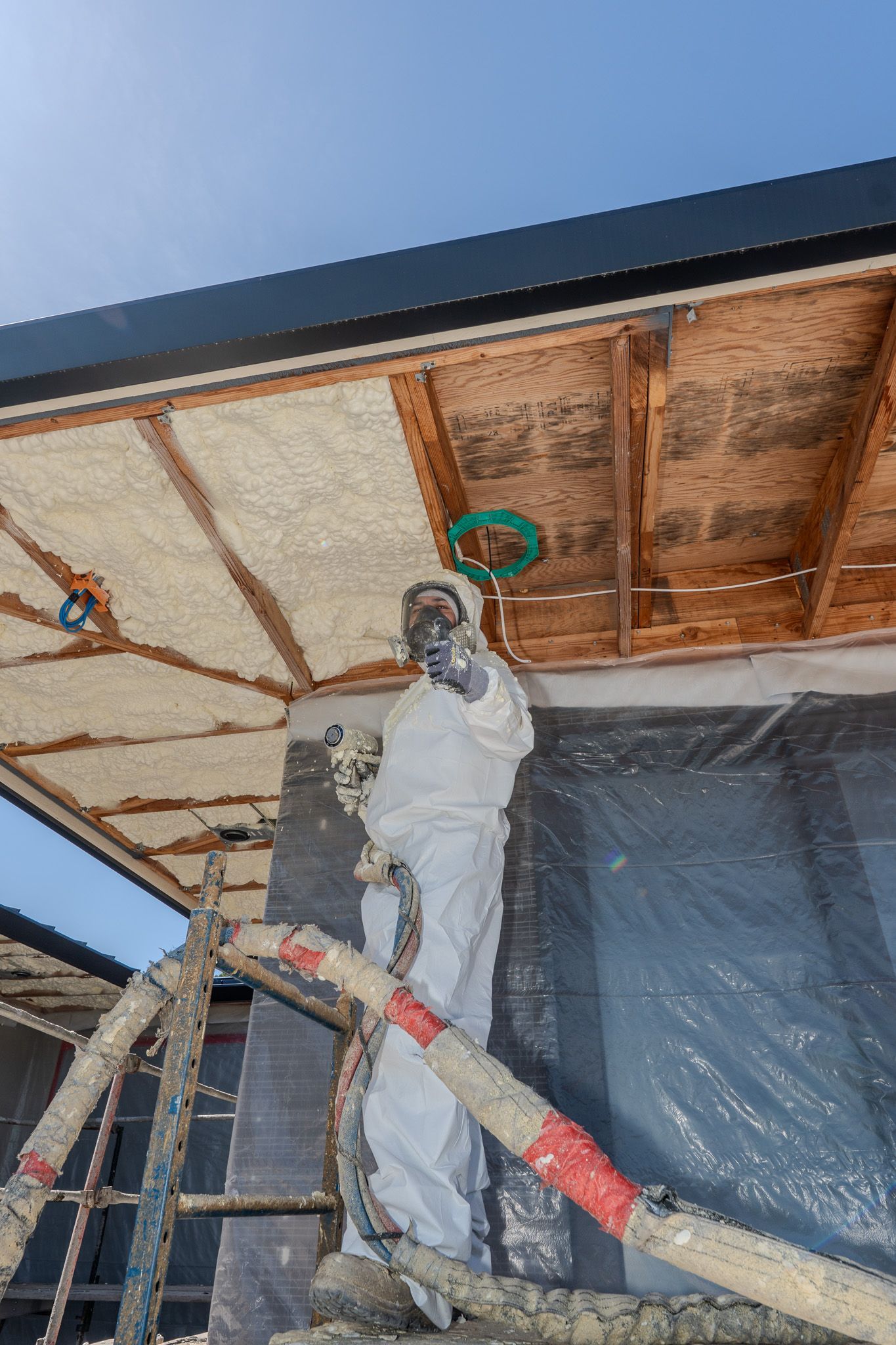 Worker painting a ceiling on a scaffold under a roof, with plastic sheeting and exposed wood beams.