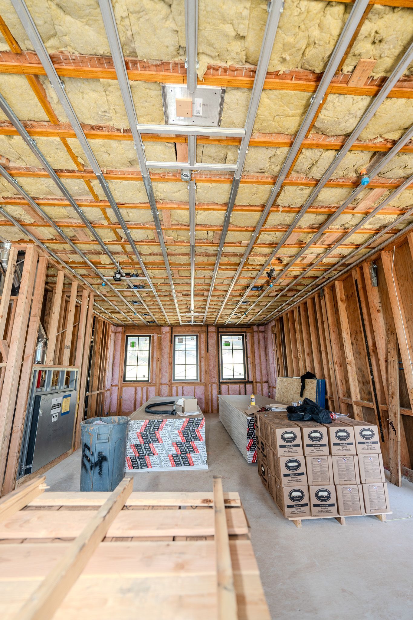 Construction site interior with exposed framing, insulation, stacked building materials, and windows at the far end