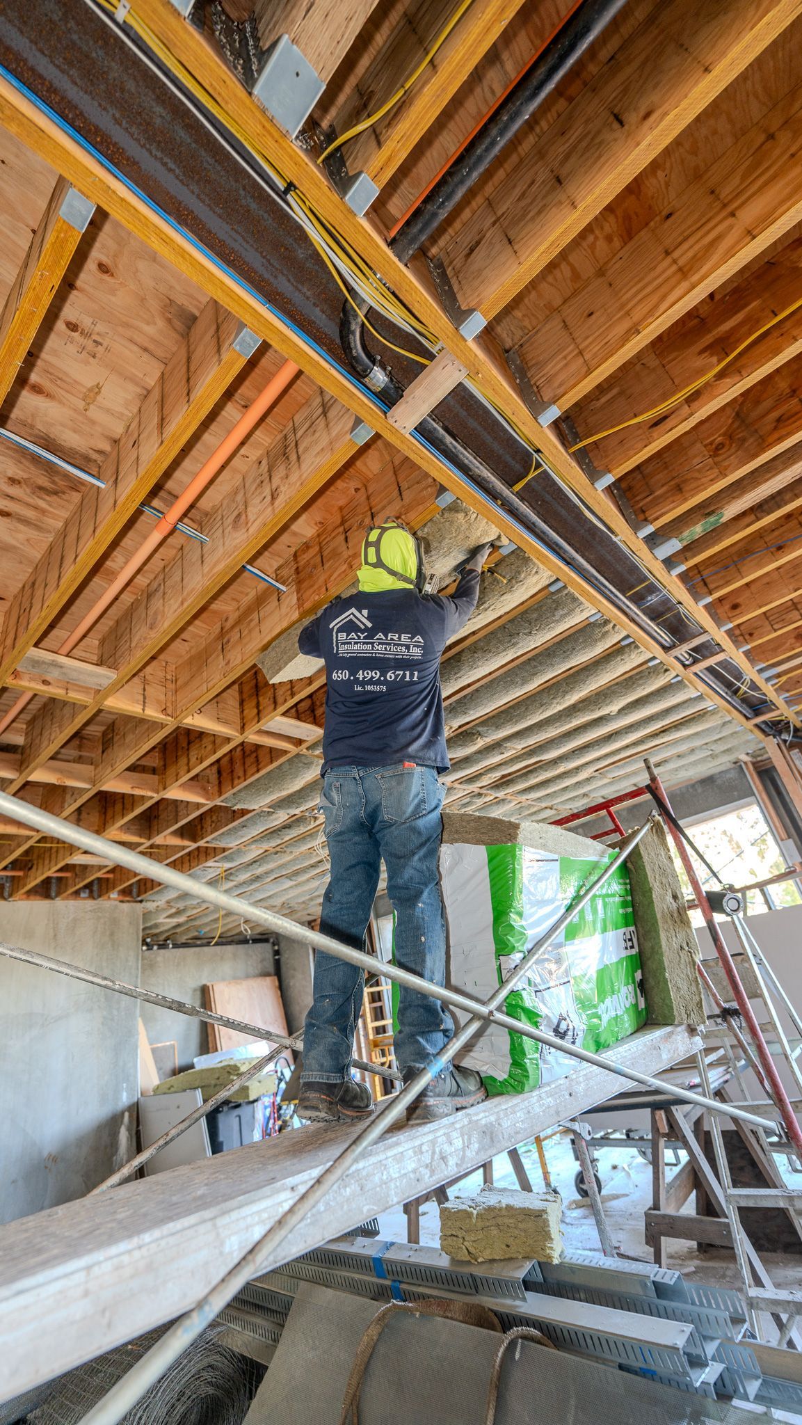 Worker insulating attic ceiling while standing on a ladder, wearing a plaid shirt and black overalls.