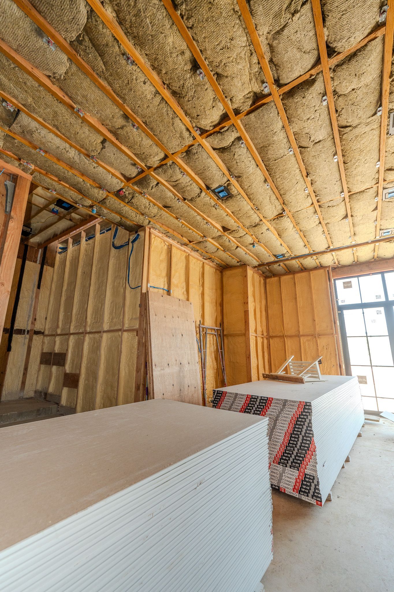 Interior of a room under construction with exposed insulation, wood framing, and stacked drywall sheets