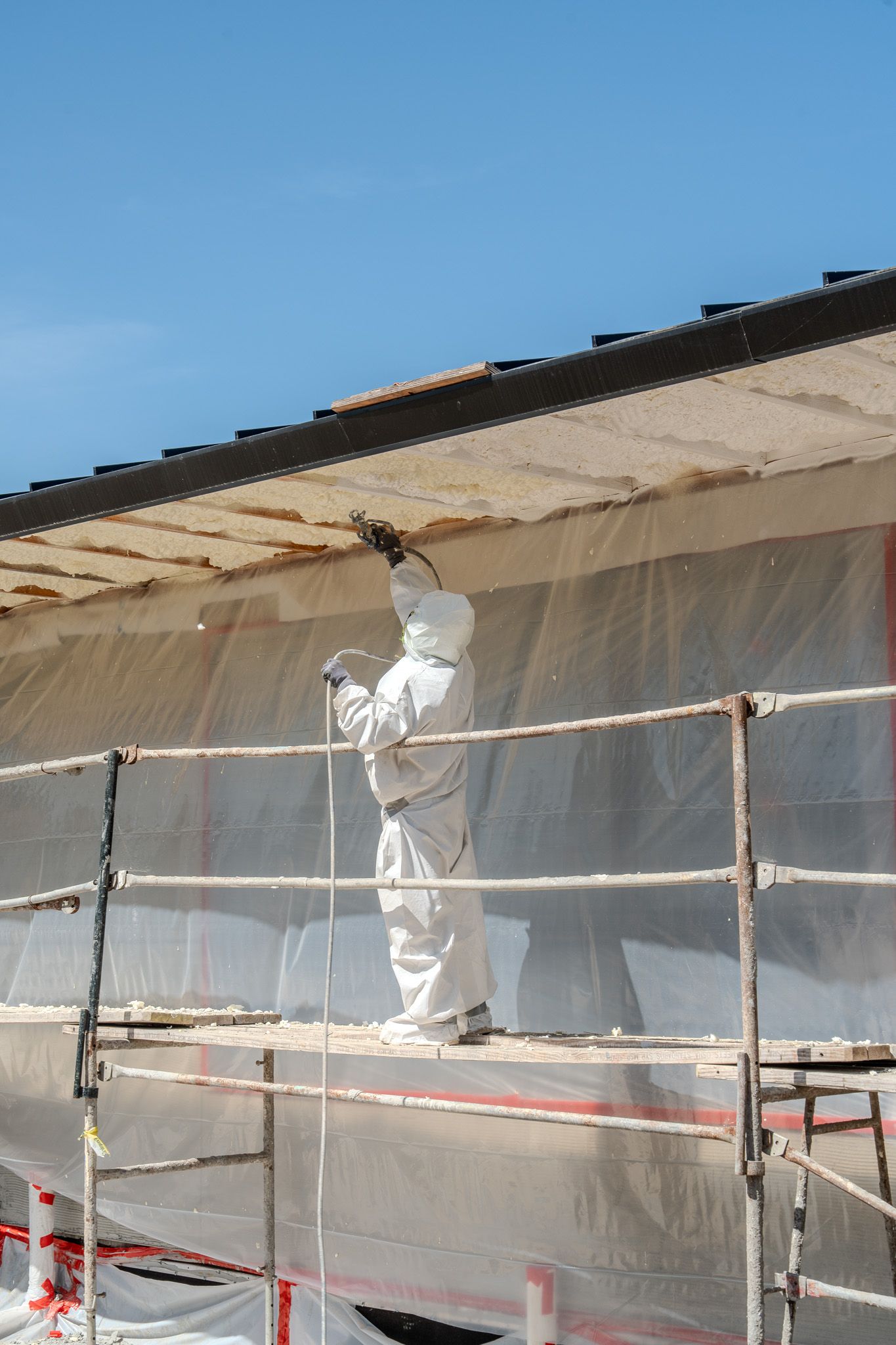 Worker in white protective suit spraying or painting a building exterior from scaffolding behind plastic sheeting