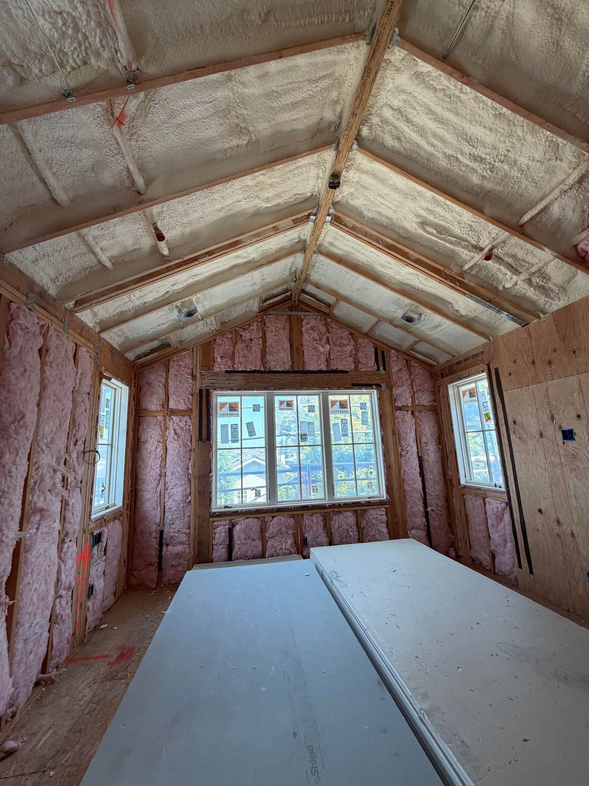 Unfinished attic room with pink insulation, exposed roof beams, and a large window at the far end.