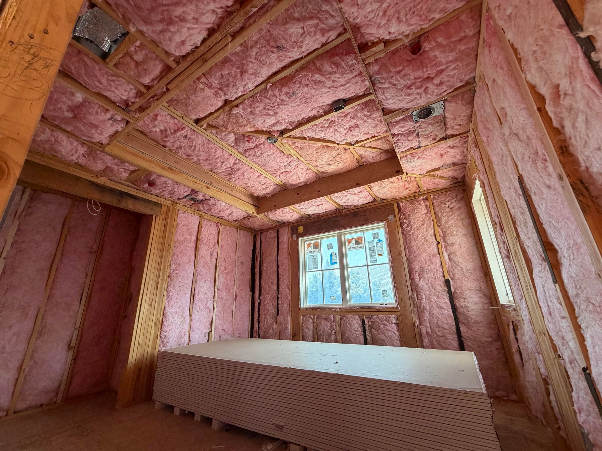 Unfinished room with pink insulation in walls and ceiling, wooden framing, and a small window.