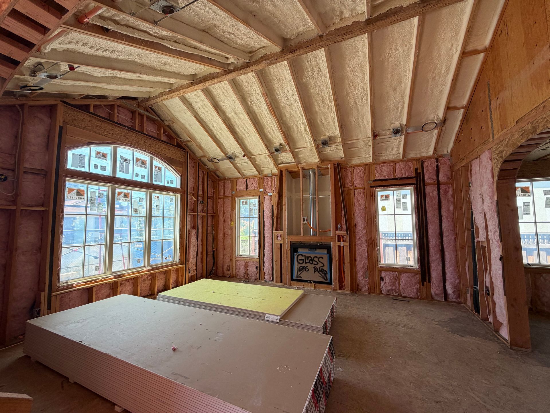 Unfinished room with exposed insulation, wooden beams, and large windows overlooking a snowy view.