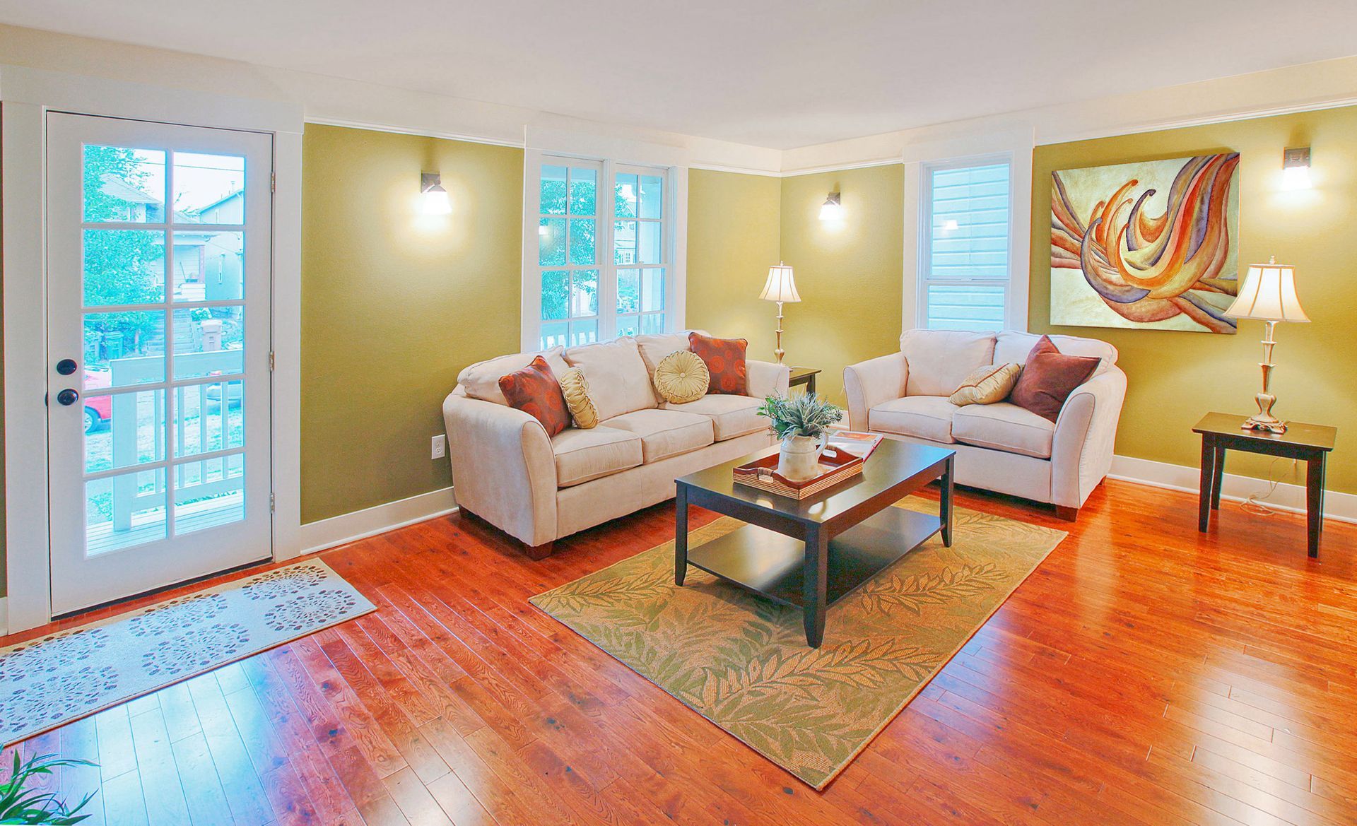 Bright living room with white sofas, orange rug, glass coffee table, and wall art by large windows.