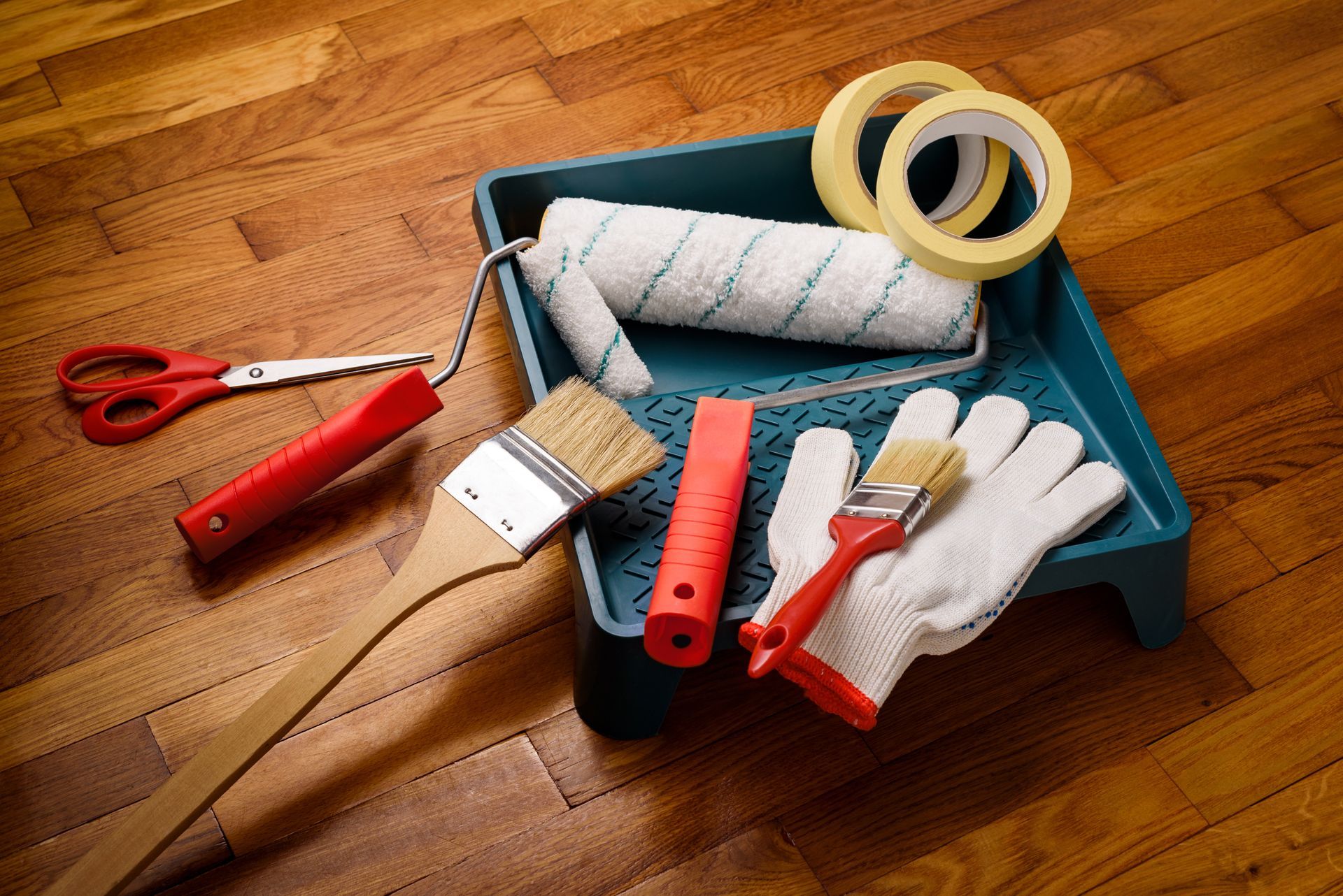 Painting tools in a blue tray on a wooden floor, including brushes, rollers, tape, gloves, and scissors.