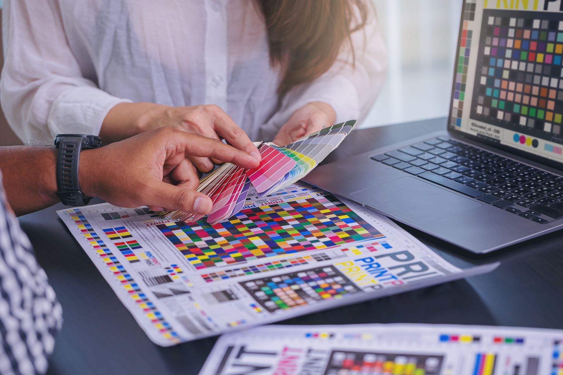 Hands reviewing colorful print swatches beside a laptop on a table.