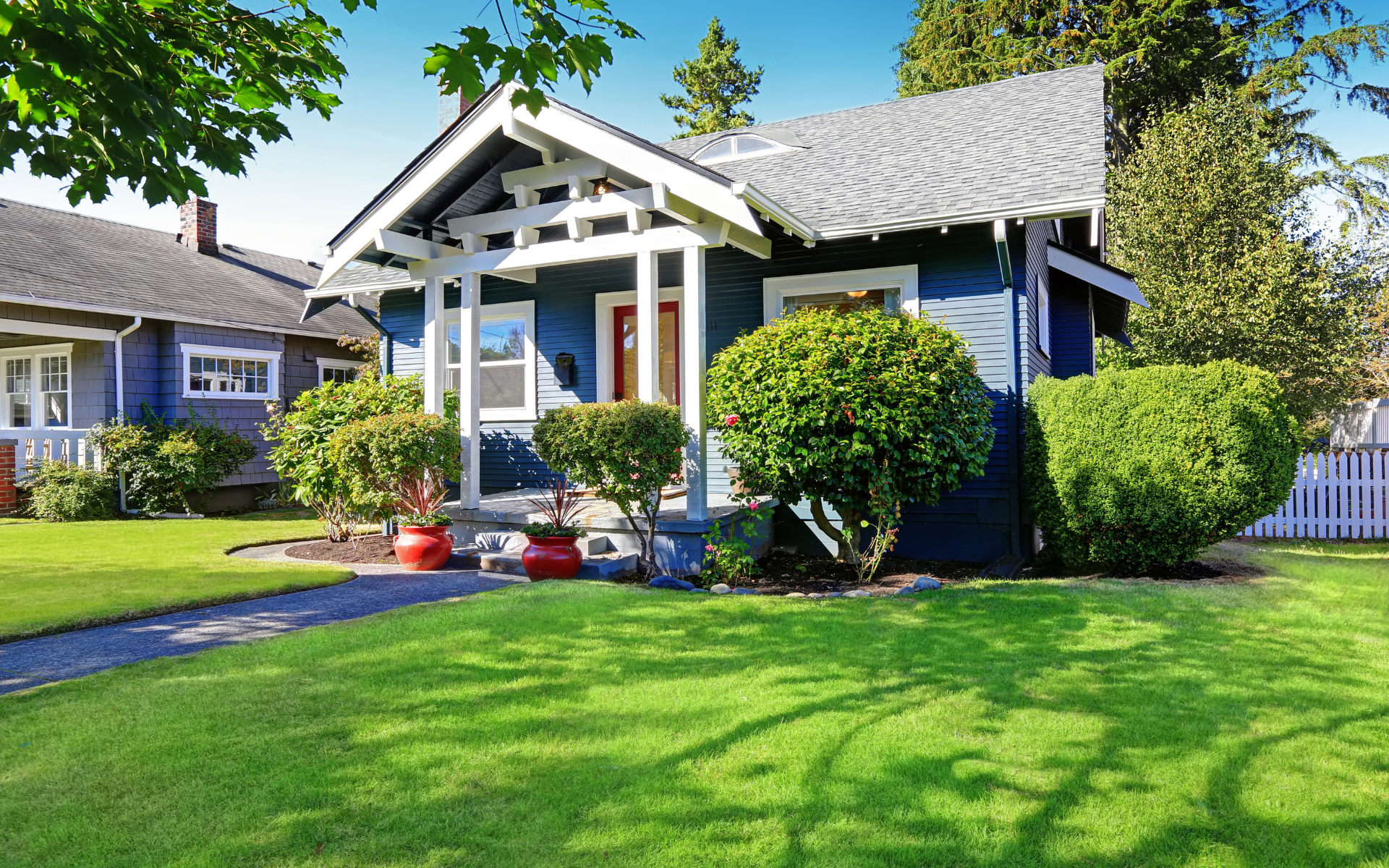 A blue craftsman-style house with a white front porch, surrounded by green lawns, shrubs, and trees under a clear blue sky.