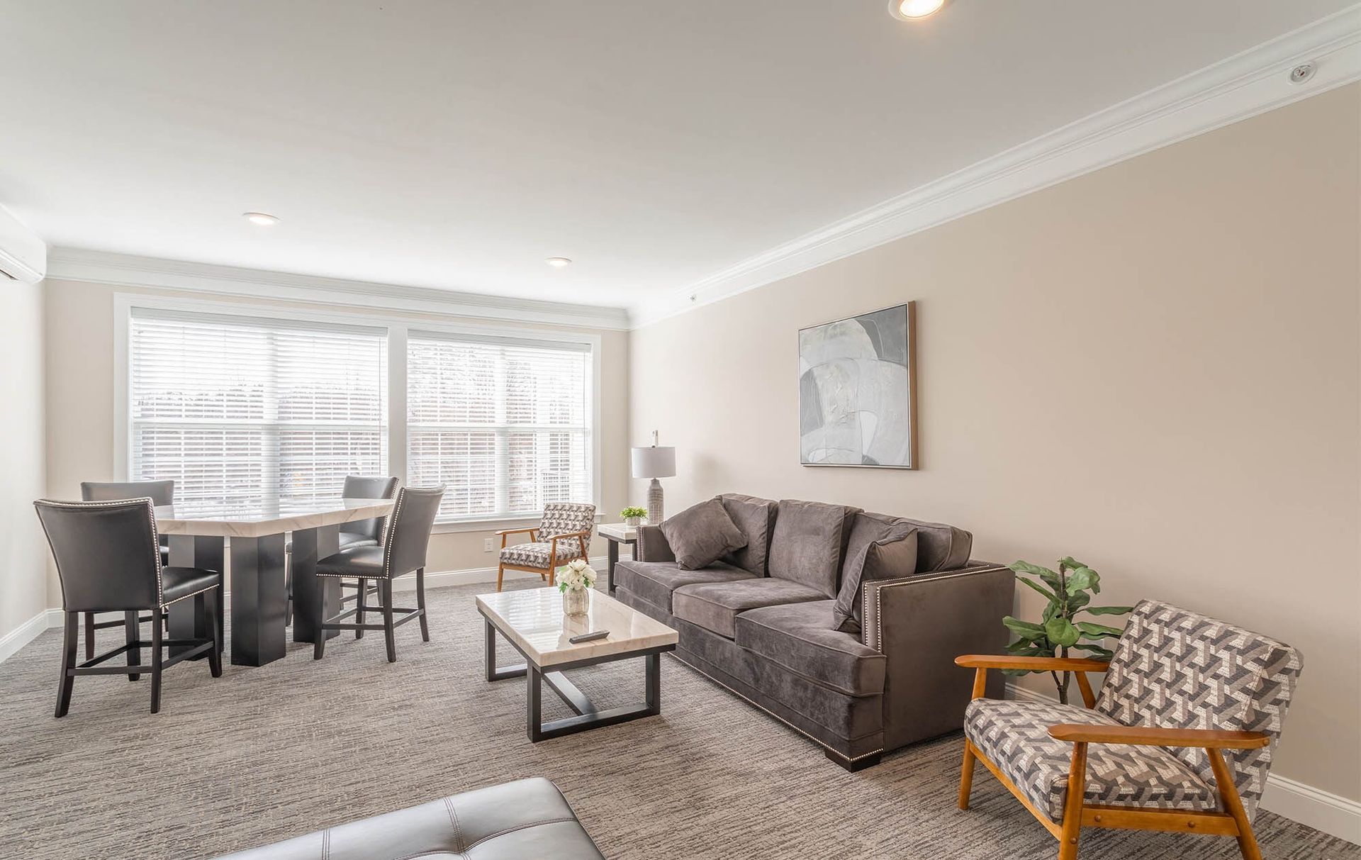 Resident lounge with beige walls, grey sofa, and large windows at The Residences at St. Joseph's Court in Levittown, PA.