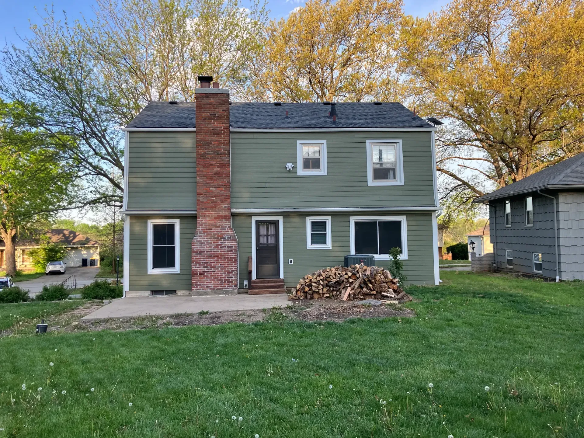 Two-story green house with brick chimney, firewood pile, and green lawn.