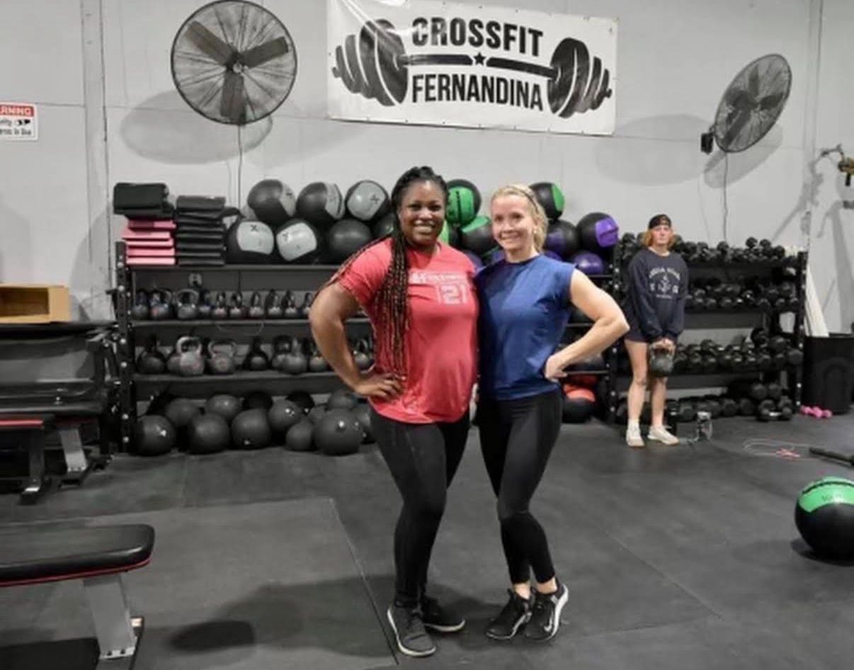 Two people posing in CrossFit Fernandina in Yulee,FL with dumbbells, kettlebells, and wall fans in the background.