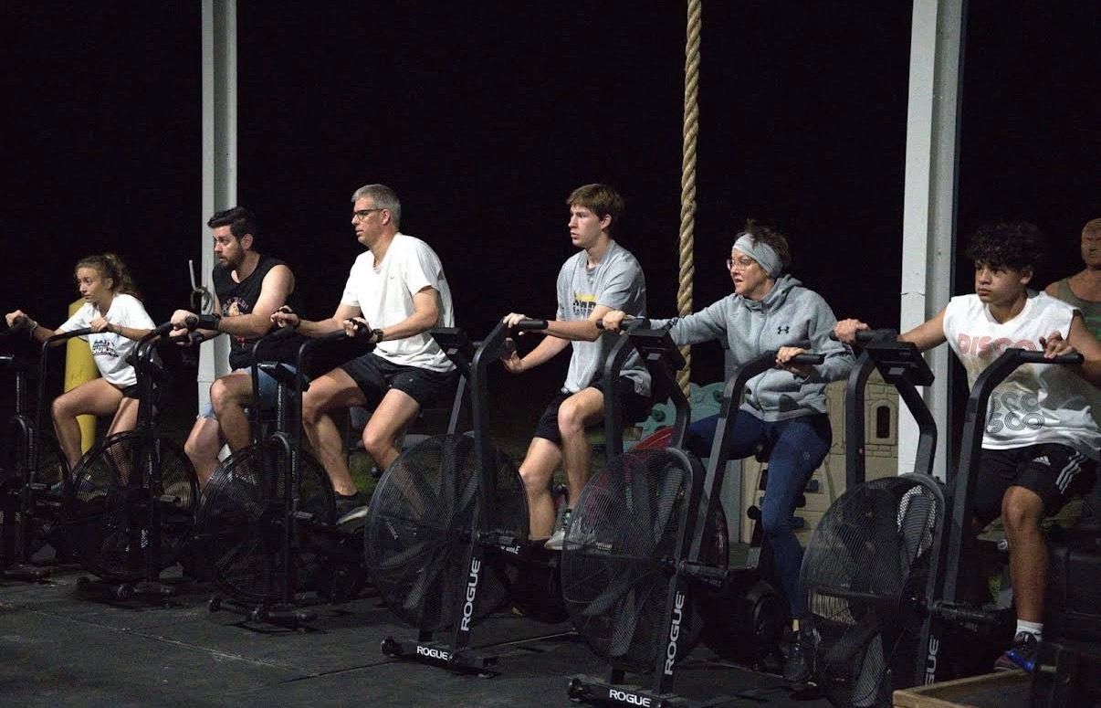 Six people pedaling stationary bikes in a dark indoor studio in CrossFit Fernandina in Yulee,FL, each holding handlebars in a row