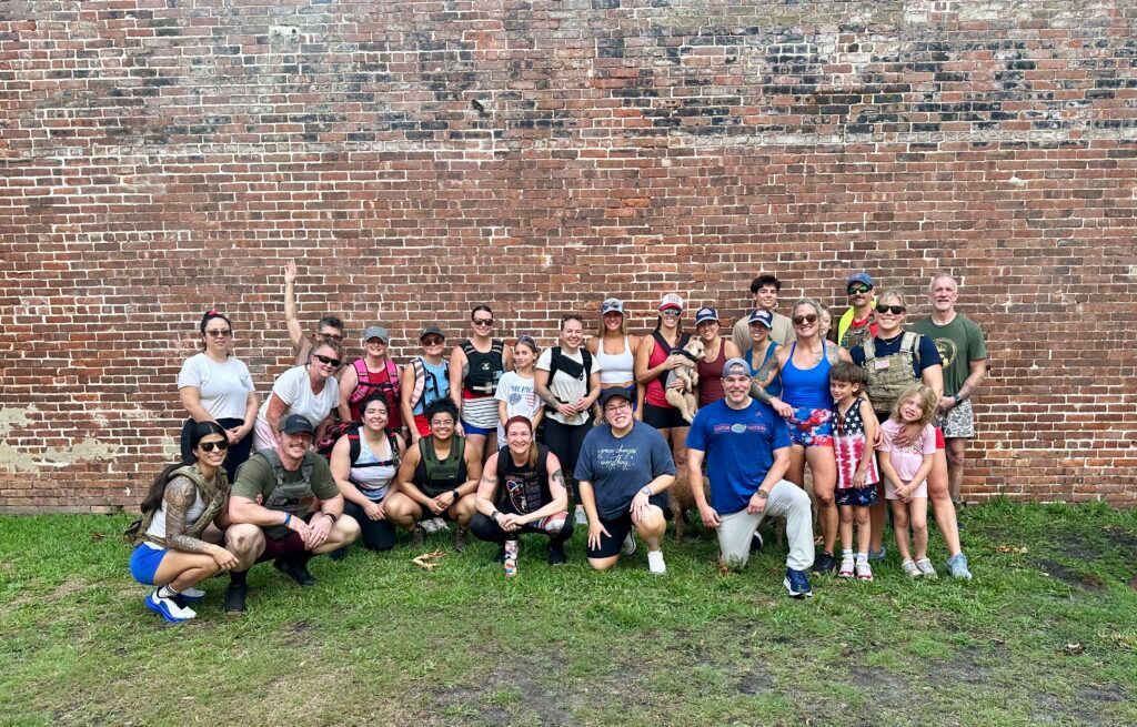 Large group posing on grass in front of a brick wall, some seated and some standing, smiling at the camera