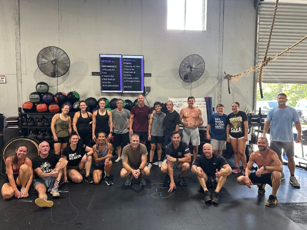 Group fitness class posing together in CrossFit Fernandina in Yulee,FL with black mats, fans, and a workout scoreboard.