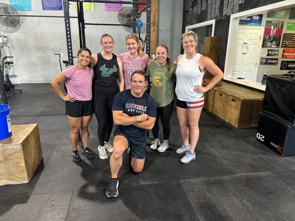 Five people posing in CrossFit Fernandina in Yulee,FL, smiling on a black rubber floor with workout equipment around them.