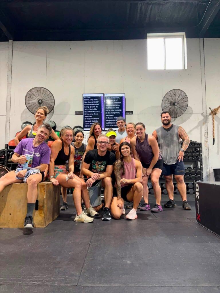 Group of people posing together in CrossFit Fernandina in Yulee,FL, seated and standing in front of wall fans and scoreboards.