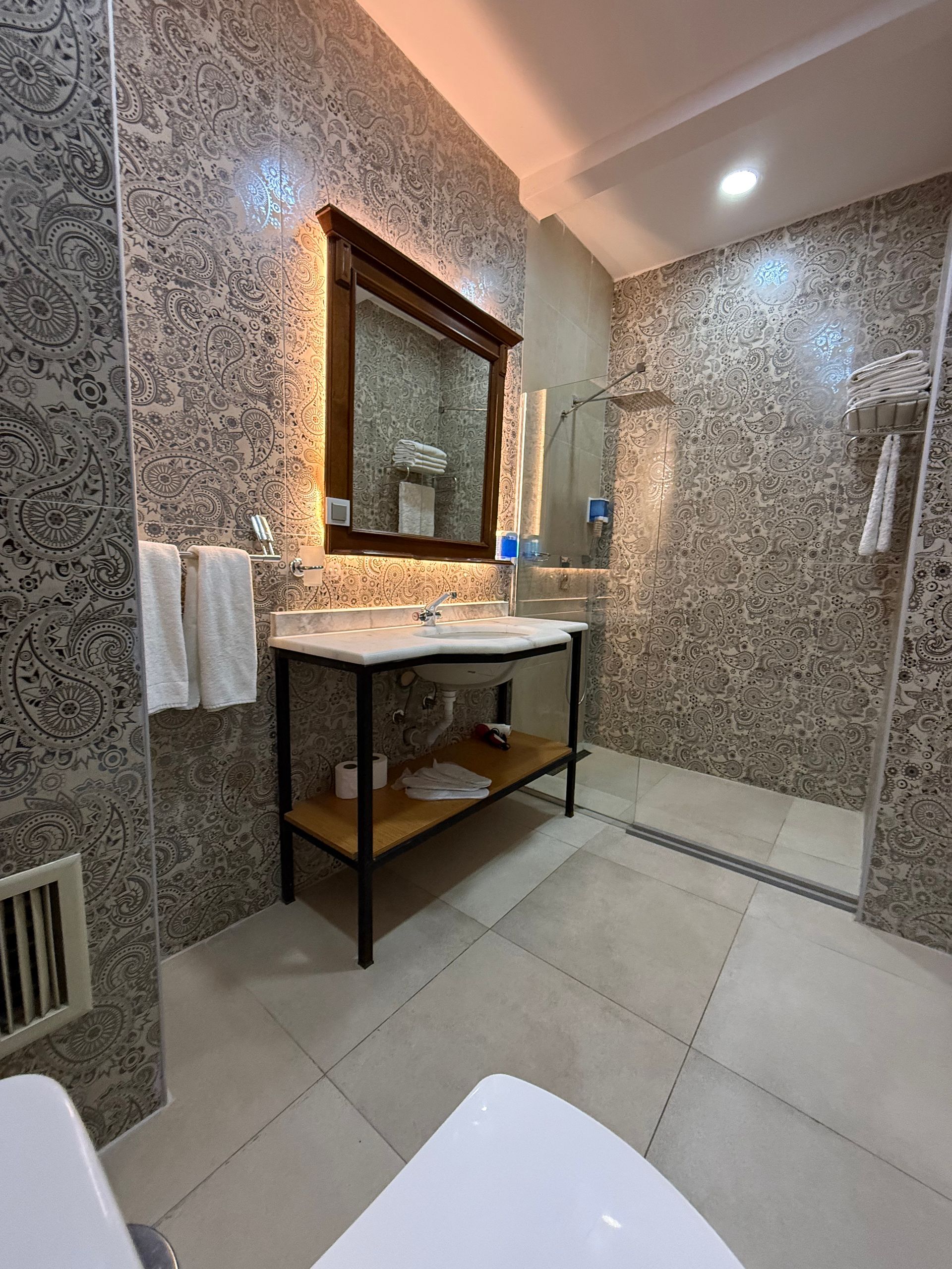 A modern bathroom with patterned tiled walls, a wooden vanity with a mirror, and a walk-in shower with light tile flooring.