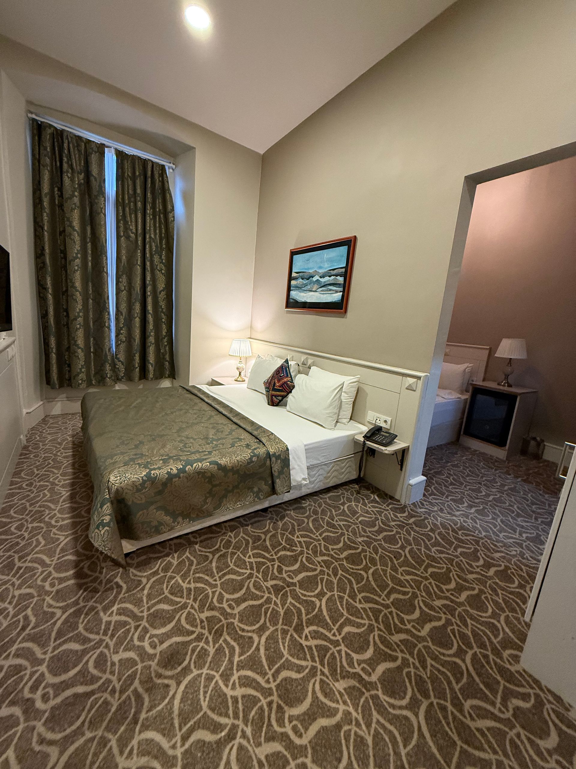 A hotel suite featuring a queen bed with patterned bedding, neutral walls, patterned carpet, and an adjacent room view.