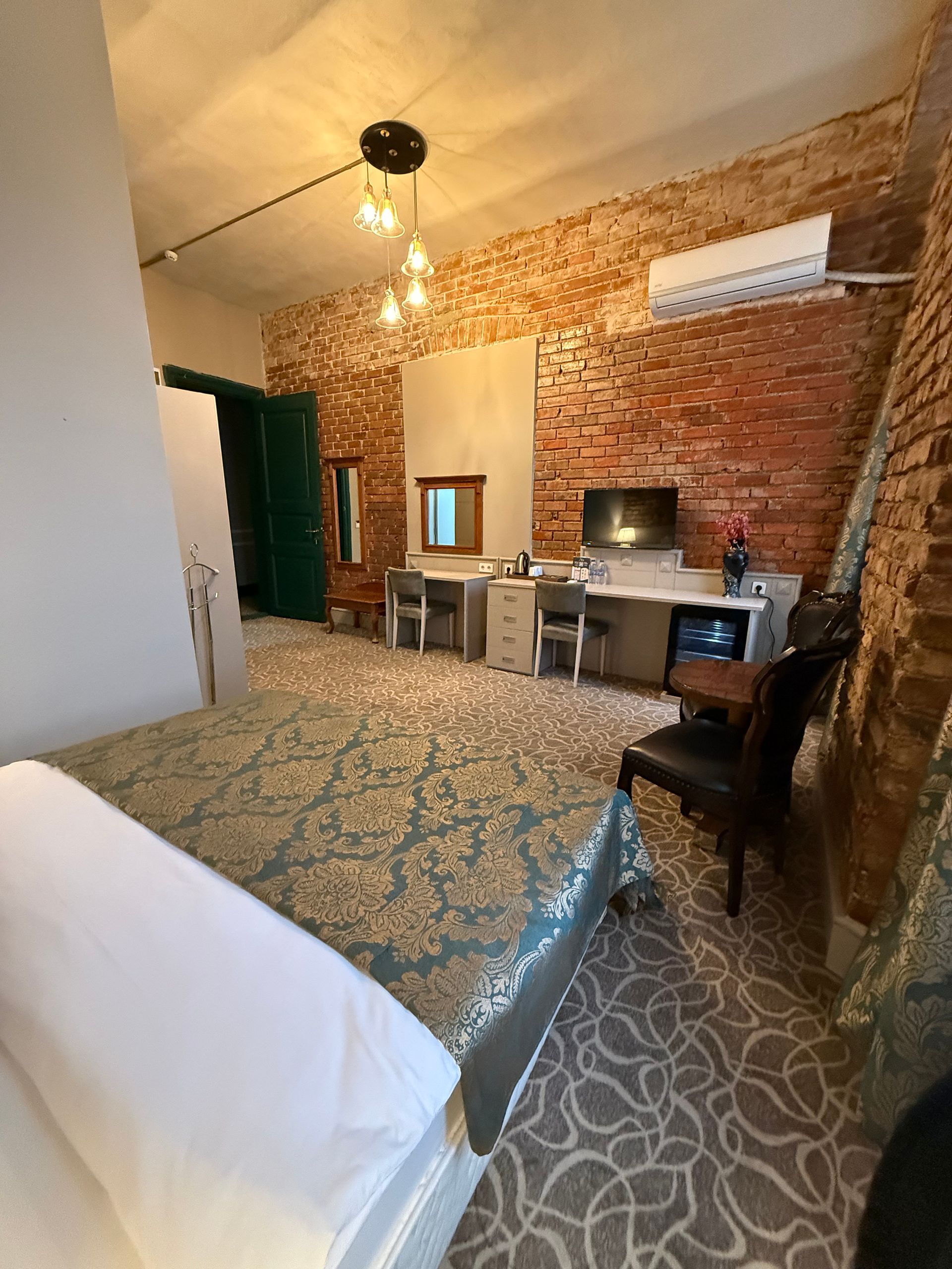 A hotel room with a patterned bedspread, an exposed brick wall, a desk with chairs, and a small TV.