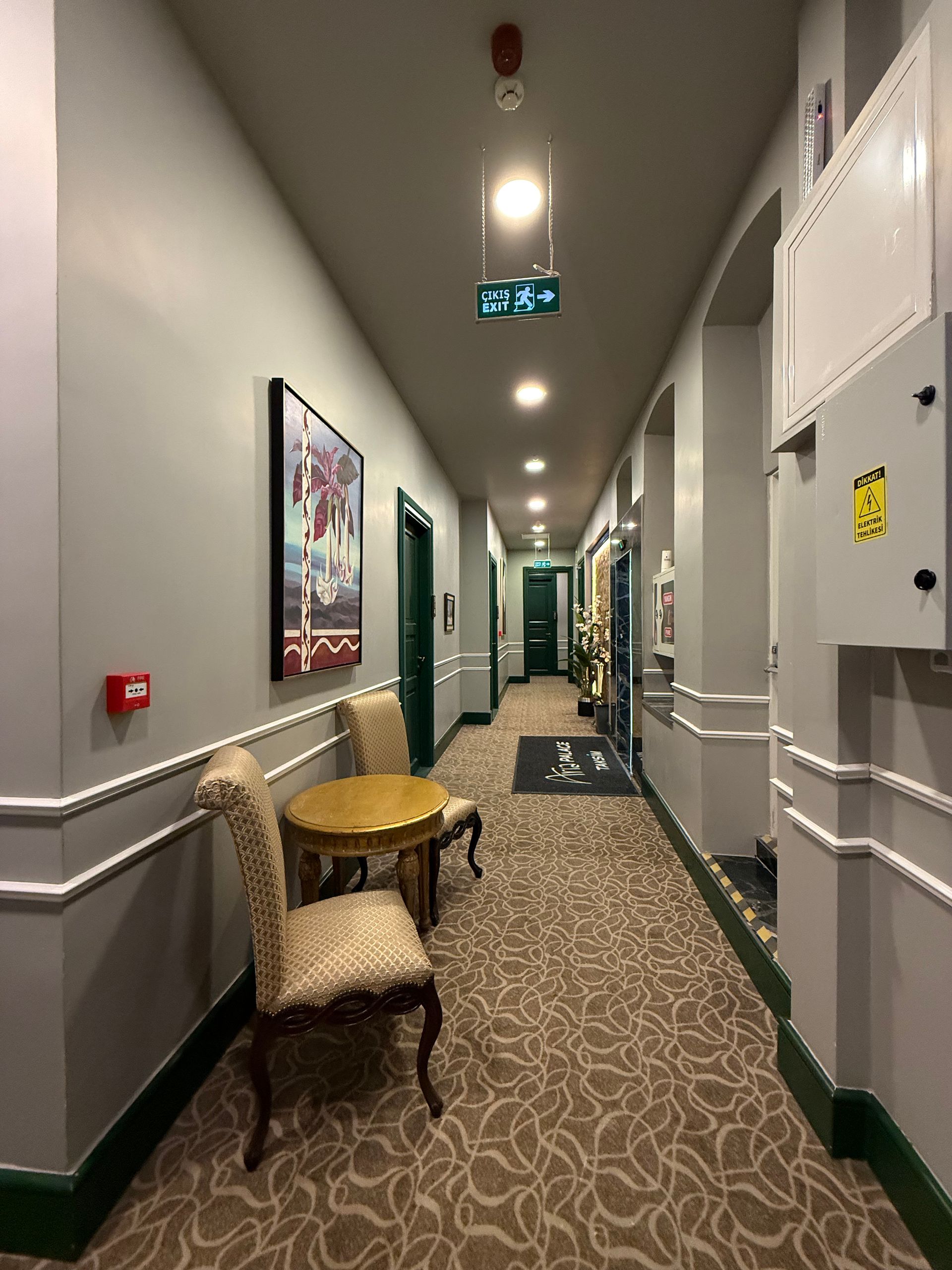 A narrow hotel hallway with patterned carpeting, light gray walls, a small table with two chairs, and an illuminated sign.