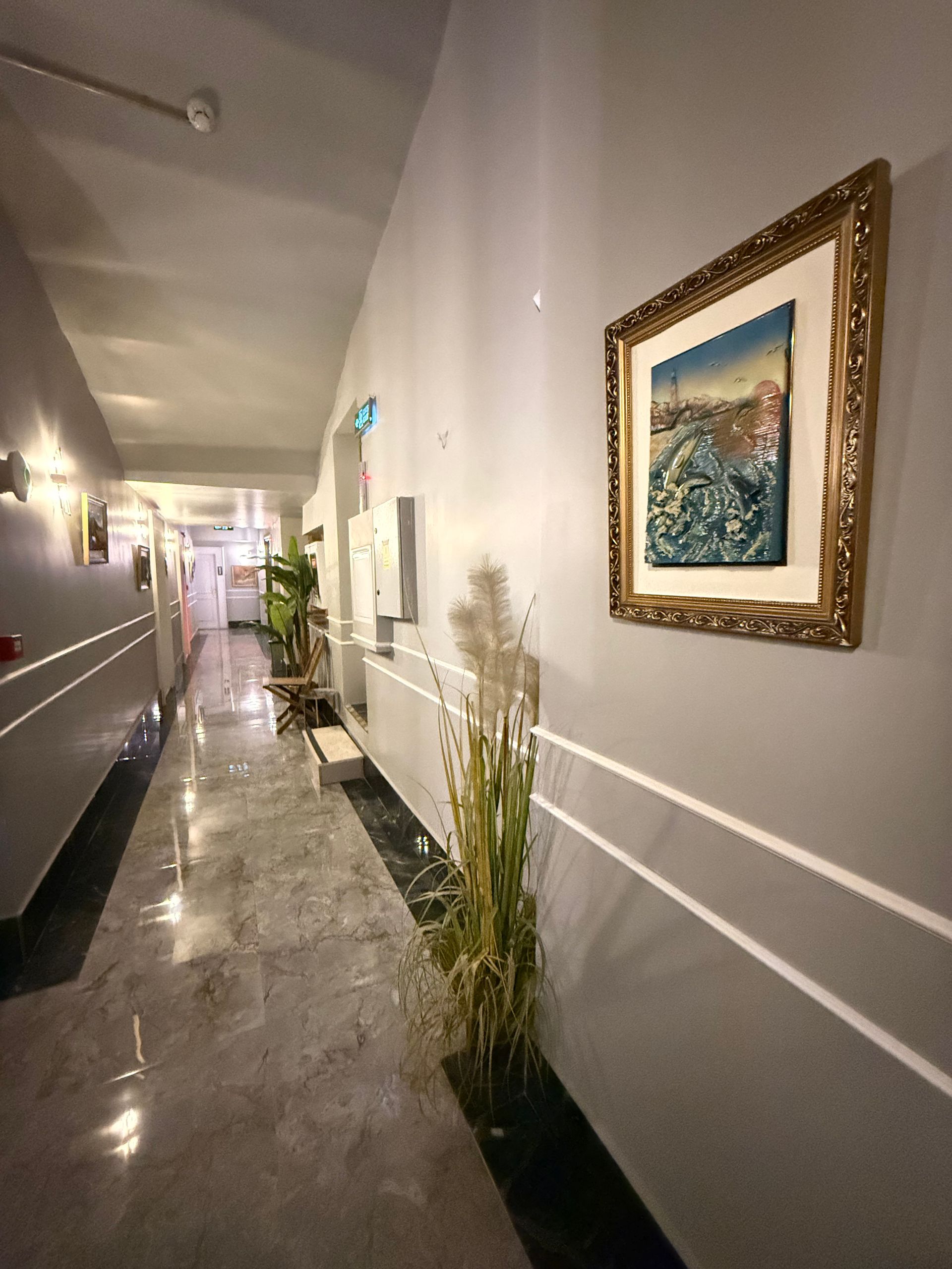 A long, narrow hotel corridor with grey walls, marble flooring, decorative plants, and a framed painting on the wall.