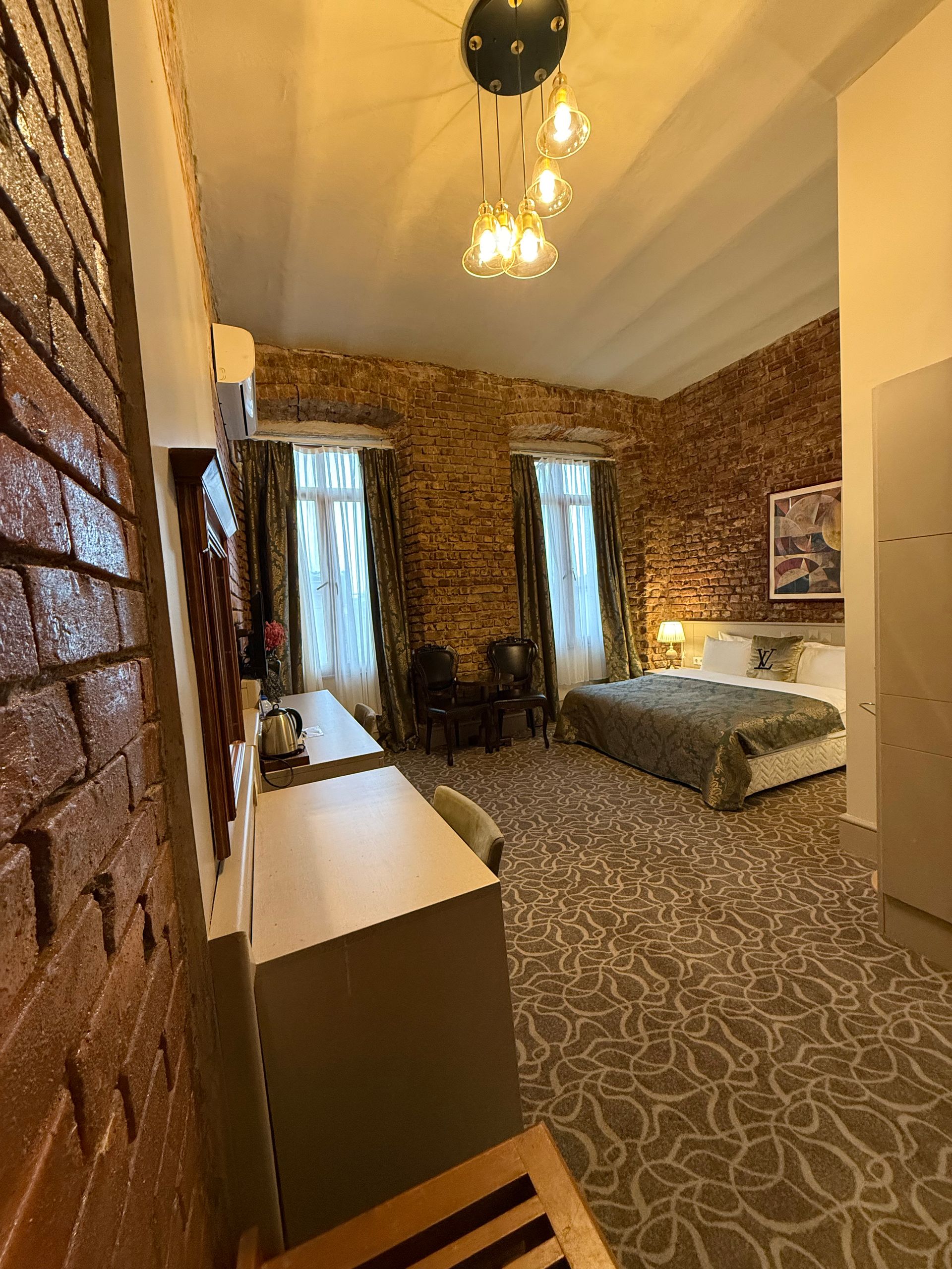 A modern hotel room with exposed brick walls, two large windows, a double bed, and a long desk with chairs.