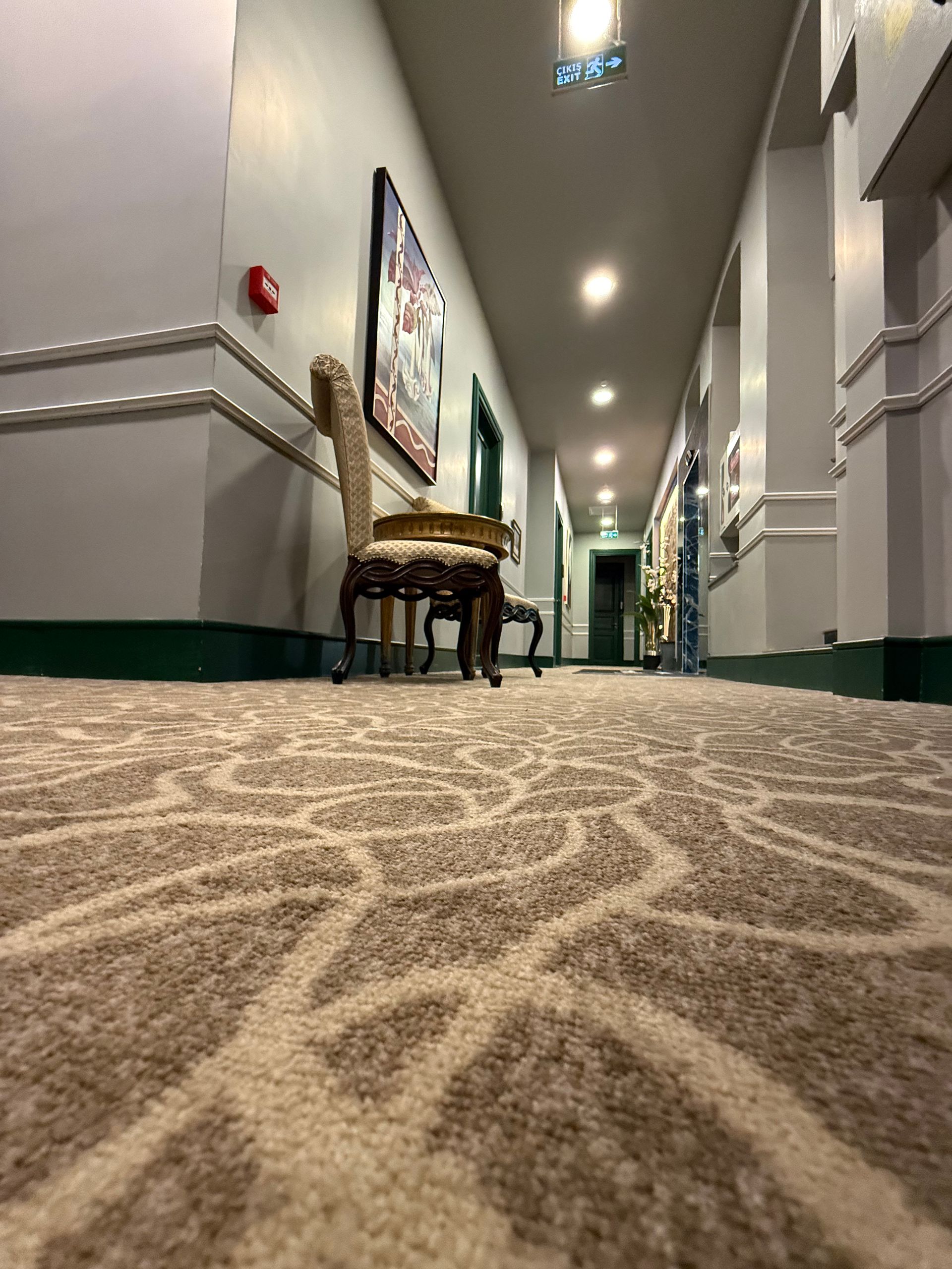 A low-angle view of a hotel hallway with light-patterned carpet, a decorative chair, and recessed ceiling lighting.