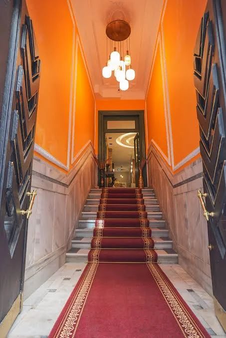 A staircase with a red runner leads up to an open doorway in a bright orange hallway with a modern hanging chandelier.