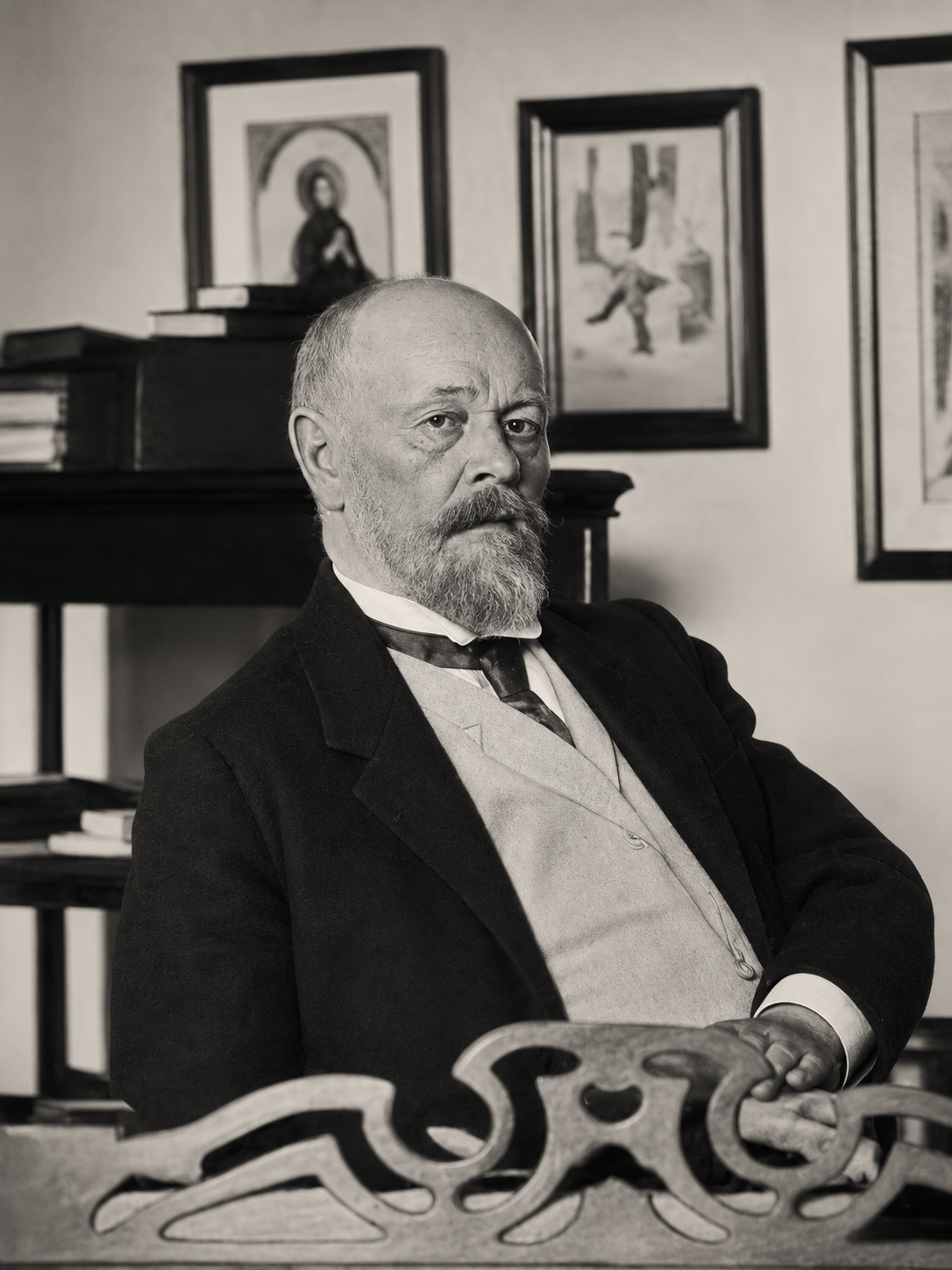 A formal, black-and-white portrait of a man with a beard and glasses sitting in a room with framed artwork on the wall.