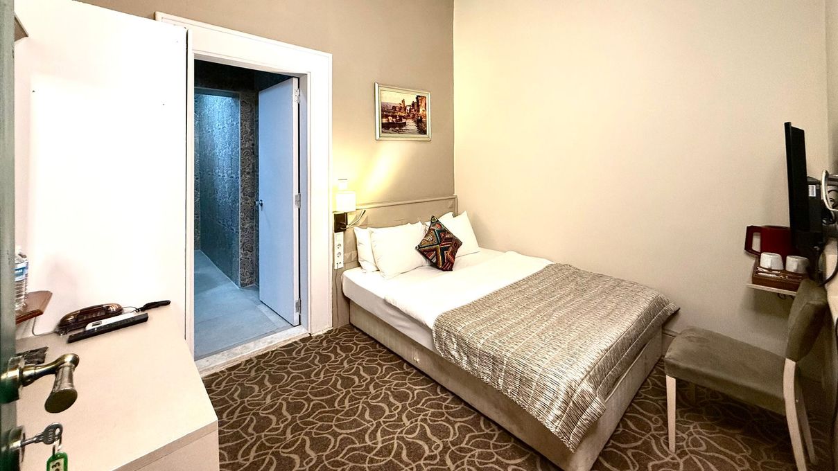 A hotel room featuring a bed with a green patterned cover, white pillows, a textured brick headboard, and a wall painting.