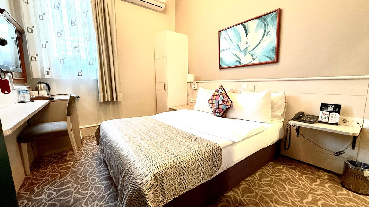 A hotel room with a white bed, patterned throw, red carpet, white curtains, a small refrigerator, and an air conditioner.